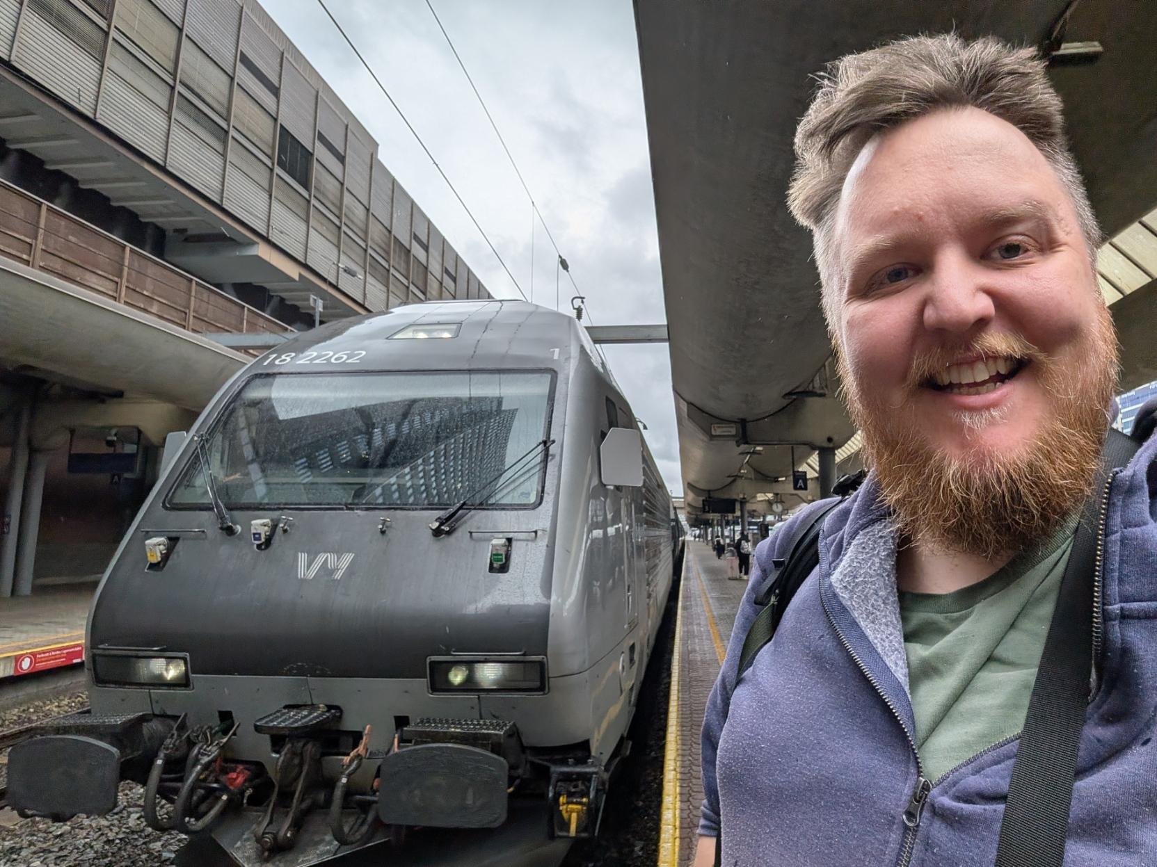 Ein selfie av ein breitt smilande mann med skjegg og lilla hettegenser på ein overbygd togperrong på Oslo Sentralstasjon. Til venstre står fronten på eit mørkegrått elektrisk lokomotiv av typen El 18 (Vy litra El 18) på sporet, klart til avgang. Lokomotivet er merka med Vy sin kvite logo på snuten og har lokomotivnummeret «18 2262» øvst over frontruta. I bakgrunnen ser ein stasjonsarkitekturen med dei karakteristiske betong- og trebygningane over spora, perrongen som strekkjer seg bakover under taket, og ein grå og overskya himmel.