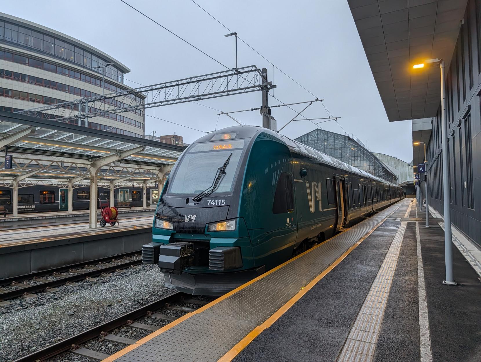 Eit mørkegrønt regiontog frå Vy, av modellen Stadler FLIRT (Type 74, settnummer 74115), står parkert ved ein perrong på ein jernbanestasjon. Toget er merka med F4 og destinasjonen Oslo S på det digitale skiltet over frontruta. Det er ein overskya og grå dag, og perrongen og spora ser våte ut. Til venstre i biletet kan ein sjå delar av stasjonsarkitekturen med overbygde perrongar og ein moderne kontorbygning i bakgrunnen.