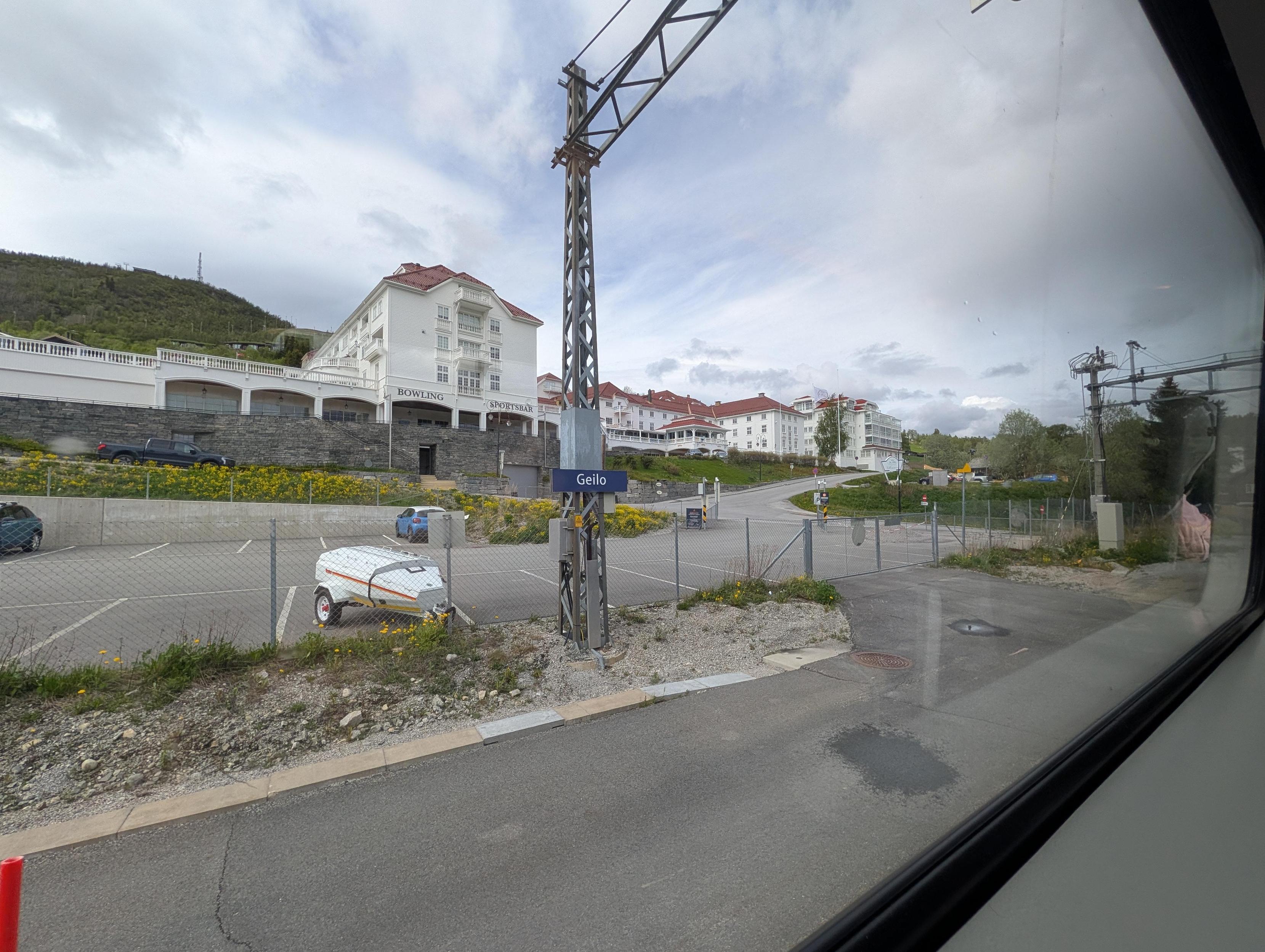 Utsyn gjennom eit togvindauge som viser Geilo stasjon og det ikoniske Vestlia Resort i bakgrunnen. Sentralt i biletet står ein mast for køyreledningar med eit mørkeblått stasjonsskilt der det står «Geilo» med kvit skrift. Bak perrongområdet og eit fletta nettinggjerde ser ein ein parkeringsplass der ein kvit tilhengar og eit par bilar står parkerte. Ein slak veg fører oppover mot det store, kvite hotellkomplekset i sveitserstil med raude tak. På fasaden til den næraste hotellbygningen står det «BOWLING» og «SPORTSBAR». Landskapet stig oppover i ein grøn, skogkledd ås under ein skyete, lyseblå himmel.