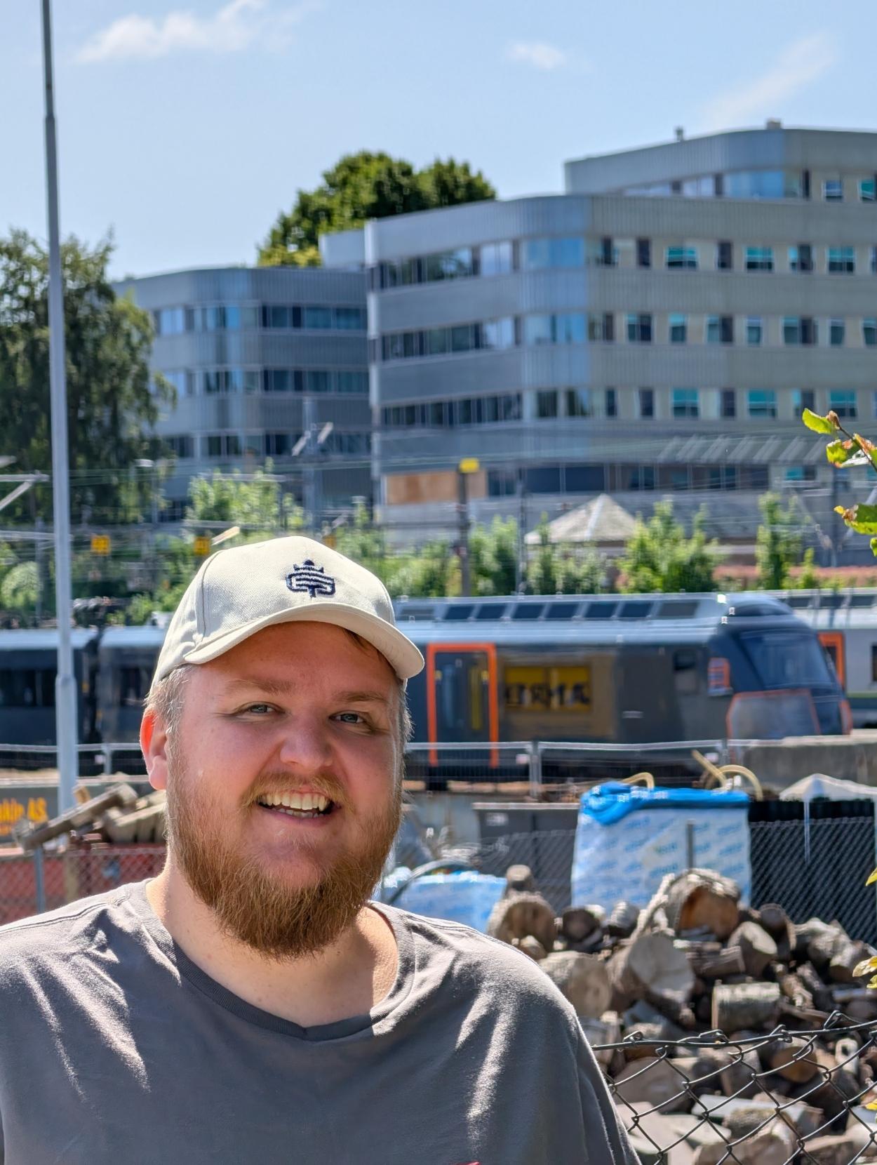 Ein selfie av ein smilande mann med skjegg, ljos caps og grå t-skjorte i framgrunnen under ein klår blå himmel. I bakgrunnen, bak eit nettinggjerde med byggeplassmateriell og ein stor stabel med vedkubbar, står eit togsett på eit spor. Toget er ein midtlivsoppgradert versjon av eit NSB Type 72 elektrisk motorvognsett frå Norske tog, opphavleg designa av Pininfarina. Togsettet er heilsvart eller mørkegrått med markerte, oransje rammer rundt passasjerdørene og langs frontpartiet. Den avrunda, aerodynamiske fronten er delvis tildekt med plast og teip. Langs heile takkanten strekkjer det seg ei tydeleg rad med skråstilde, rektangulære ventilasjonsgitter. I eit av dei store sidevindauga ser ein speglinga av den gule teksten «ARMERING» frå byggeplassen. Helt i bakgrunnen reiser det seg store, moderne kontorbygningar i sølvgrått stål og glas bak grøne tre.