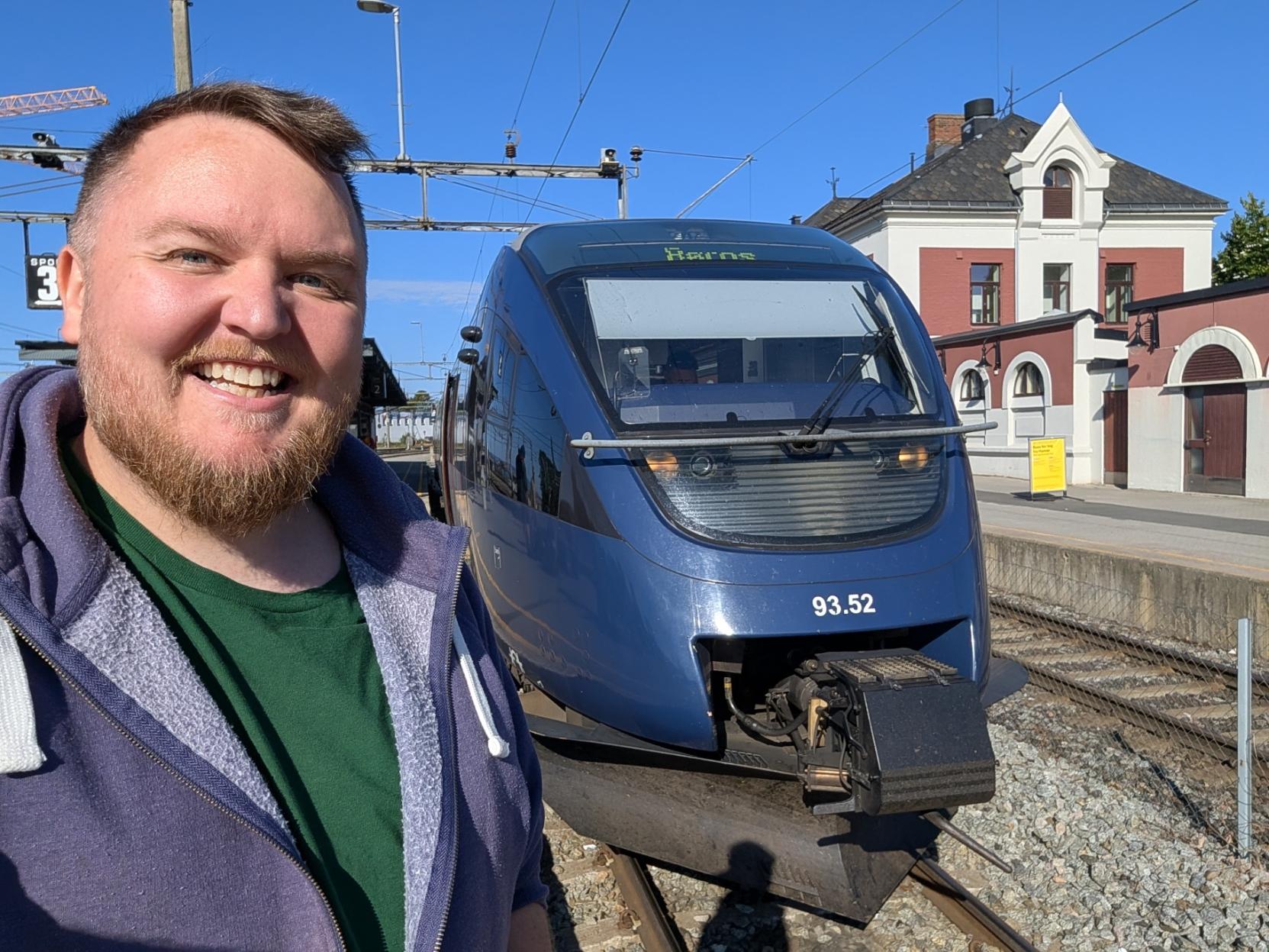 Ein selfie av ein breitt smilande mann med skjegg, grøn t-skjorte og lilla hettejakke på ein solfylt perrong på Hamar stasjon (Spor 2). Til høgre står den avrunda fronten på eit blått dieselkrengjetog av typen NSB Type 93 (SJ Nord litra BM93), klart til avgang på Rørosbanen. Under frontruta står settnummeret «93.52», og på destinasjonsskjermen over frontruta lyser «Røros» i grøne punkt. Frontlysa er tende, og vognsida ber SJ Nord-logoen og merkinga «BCM93.52». I bakgrunnen til høgre ligg den raude og kvite stasjonsbygningen med kvite gavlar og buete vindauge, og til venstre reiser ei oransje byggjekran og køyreleidningane seg mot ein klår, blå sommarhimmel.

🚆 Tog: Type 93 · Litra: BM93 · Nr: 93.52 · Stasjon: Hamar · Bane: Rørosbanen · Selskap: SJ Nord