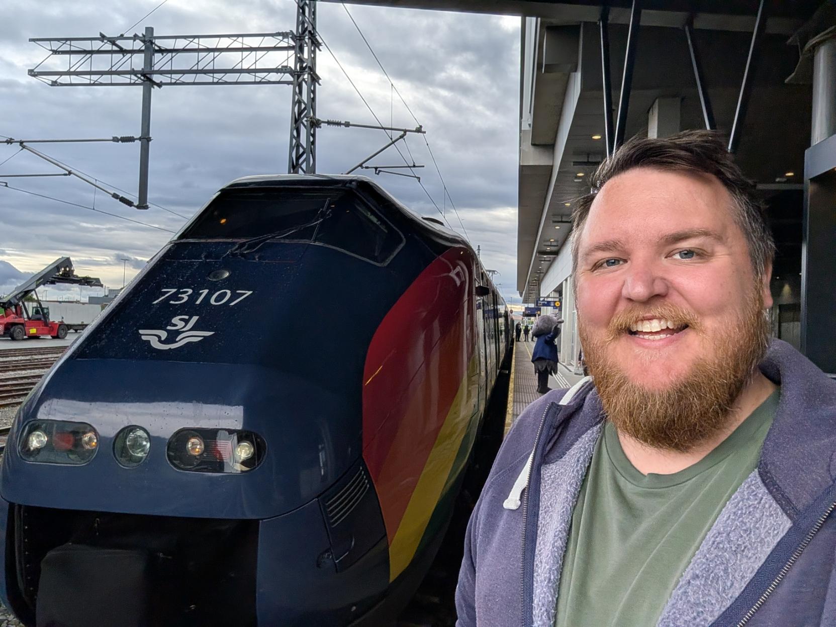 Ein selfie av ein breitt smilande mann med skjegg, grøn t-skjorte og blå hettejakke på ein perrong ved Trondheim S. Til venstre står den spisse fronten på eit mørkeblått elektrisk krengjetog av typen NSB Type 73 (SJ Nord-sett 73-07), som skal kjøre F6-ruten over Dovrebanen til Oslo S. Togfronten er merka med settnummeret «73107» og SJ Nord-logoen i kvit skrift. Langs sida av toget snor eit fargerikt mønster i raudt, gult og grønt seg utover karosseriet, og frontlysa lyser. I bakgrunnen til venstre ser ein eit godsområde med ein raud gaffeltruck under ein skya himmel, og lenger bak på perrongen står ein maskottfigur i utvalde klede.