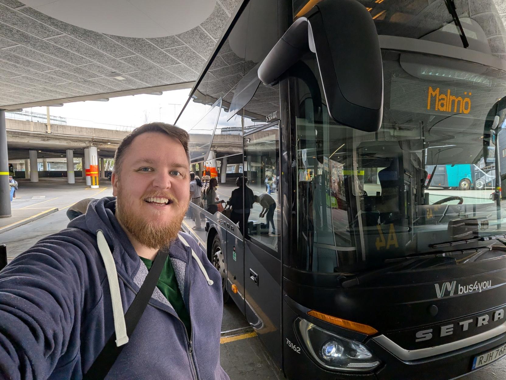Ein smilande mann med skjegg tek ein selfie på ein bussterminal under eit betongtak. Til høgre for han står ein stor, mørkegrå dobbeltdekkarbuss av merket Setra (modell S 531 DT). Bussen er merka med «Vy bus4you» på fronten, og destinasjonsskiltet lyser gult med teksten «Malmö». I bakgrunnen kan ein sjå andre reisande som gjer seg klare til å gå om bord, samt søyler og ein overbygd køyrebane på terminalområdet.