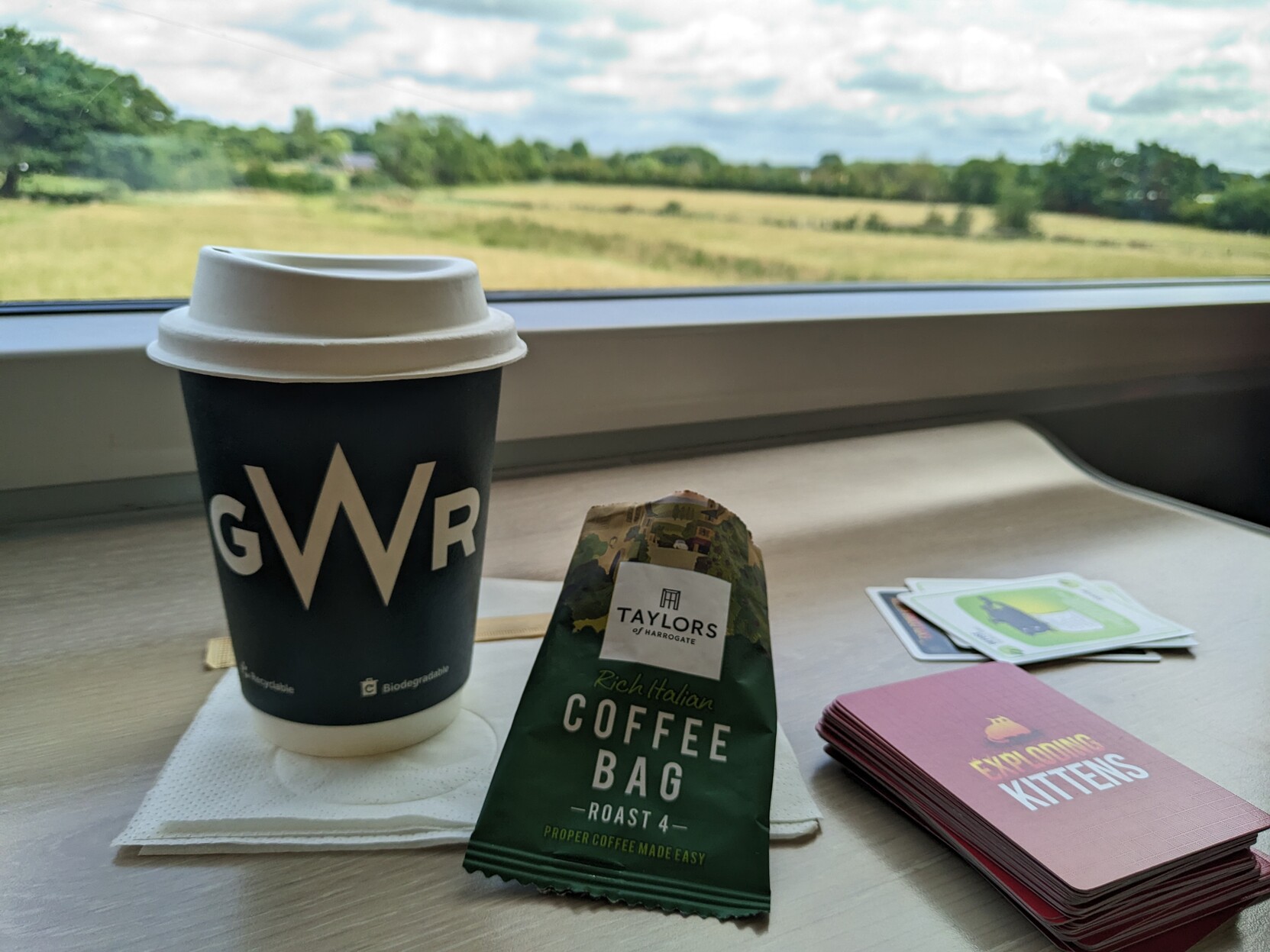 Bilete av kaffikopp med teksten "GWR" på. Ei lita, open pose merka "Taylor's Coffee Bag". På sida ligg ein kortstokk frå spelet "Exploding Kittens".

I bakgrunnen ser ein ut eit togvindauge på engelsk landskap.