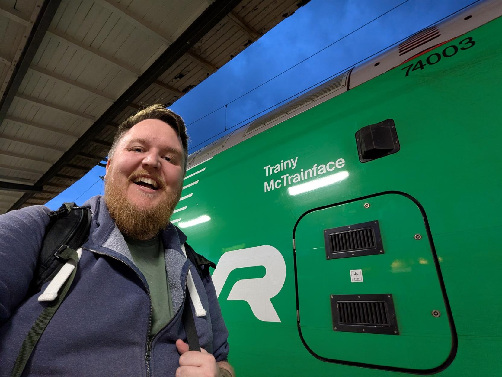 Ein selfie av ein kjempefornøgd, smilande mann med skjegg og lilla hettegenser på ein perrong om kvelden. Til høgre dominerer den skinande, knallgrøne sida på eit elektrisk snøggtog av typen Stadler FLIRT (VR litra X74). På vognsida står det legendariske tognamnet «Trainy McTrainface» skrive i kvit skrift over den kvite «VR»-logoen. Oppe til høgre ser ein vognnummeret «74003». Over toget og perrongtaket til venstre kan ein sjå ein djupt mørkeblå kveldshimmel.