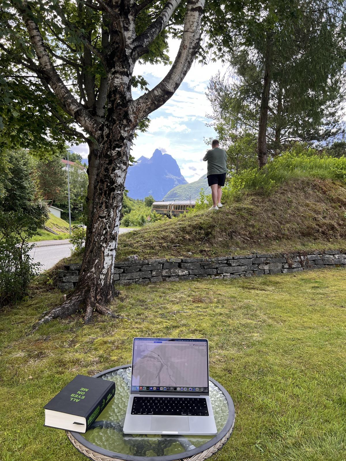 Ein open sølvfarga MacBook står på eit rundt glasbord i ein grøn hage, med Bane NOR sitt togkart på skjermen. Ved sida ligg ei tjukk innbunden bok med tittelen «All Eyes on Me». Til venstre veks ei stor bjørk med kvit-svart bork. Bak ein låg tørrmur reiser ein grasbakke seg, og oppå står ein mann i grøn t-skjorte og mørke shorts med ryggen til, vend mot jarnbana nedanfor. Der er eit lyst, gamaldags tog delvis synleg mellom trea. I bakgrunnen tårnar dei bratte Romsdalsfjella seg opp mot ein blå himmel med lette skyer, med den spisse toppen Romsdalshornet midt i biletet.