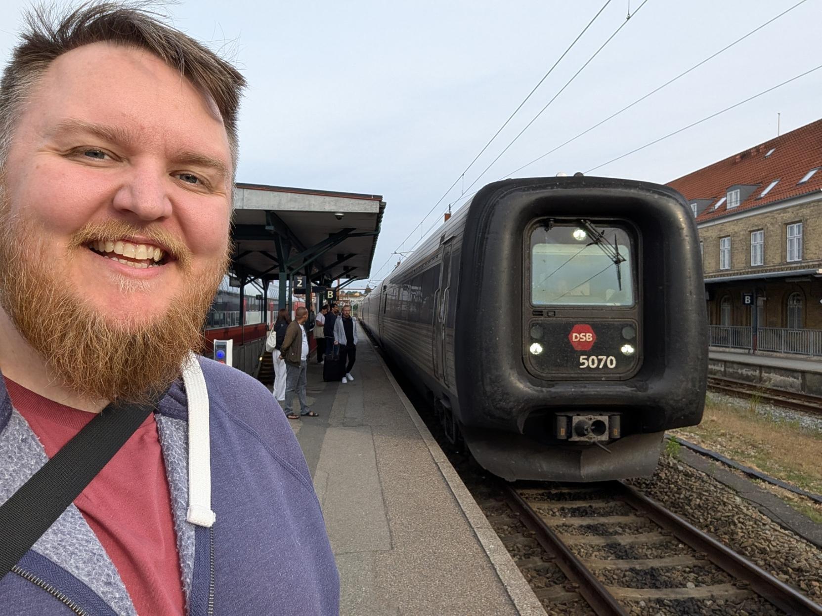 Ein selfie av ein breitt smilande mann med skjegg og lilla hettegenser på ein togperrong på Næstved stasjon. Til høgre står fronten på eit elektrisk DSB IR4-tog (vognsett 5070) på sporet, lett attkjennande på den tjukke, svarte gummimembranen som omkransar frontruta, og DSB sin raude logo på snuten. Togsettet strekkjer seg bakover langs den grå perrongen, der nokre passasjerar står oppstilt under perrongtaket til venstre. I bakgrunnen til høgre ser ein den tradisjonelle stasjonsbygningen i gulbrun murstein med raudt tak under ein ljos, klår kveldshimmel.