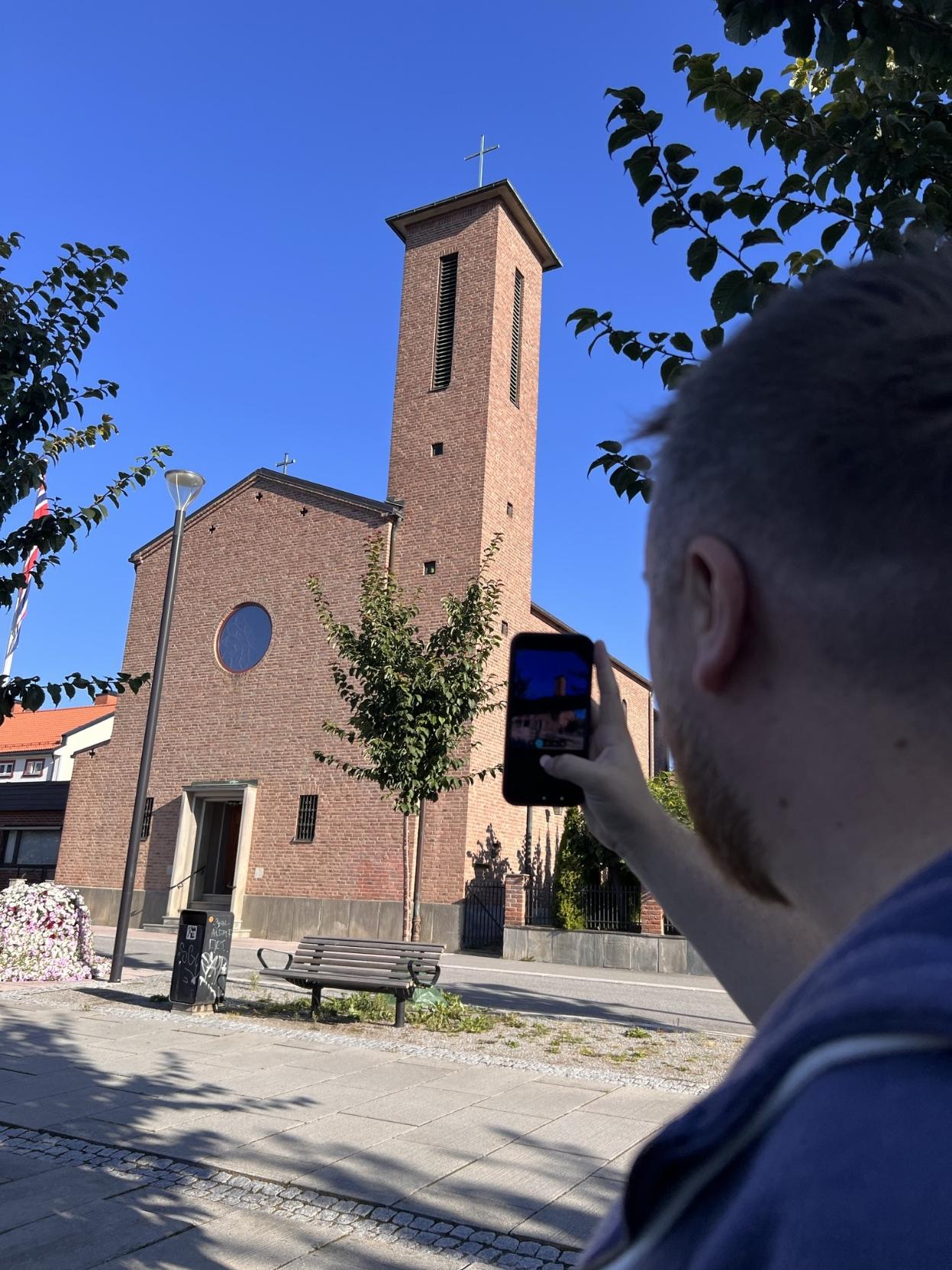 Ein mann sett bakfrå tek bilete med smarttelefonen sin av ei kyrkje bygd i raud teglstein. Kyrkja har eit høgt, slankt tårn med ein kross på toppen og eit rundt vindauge på fasaden. Framføre kyrkja står ein trebenk og eit lite tre under ein klår, blå himmel.