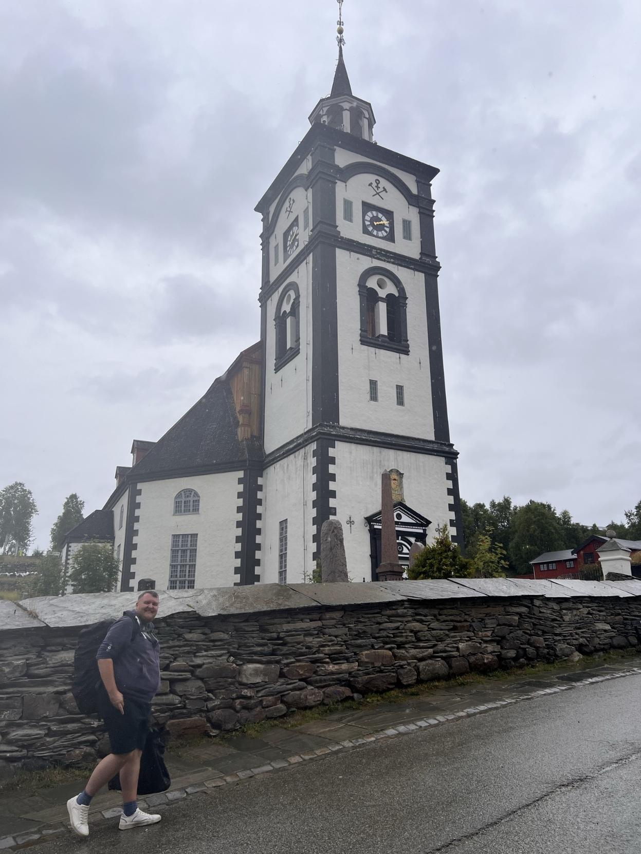 Ein smilande mann med stor ryggsekk går langs ein våt veg i overskya vêr. Bak ein låg skifermur ruvar Røros kyrkje med sine karakteristiske kvite murar og mørke detaljar.
