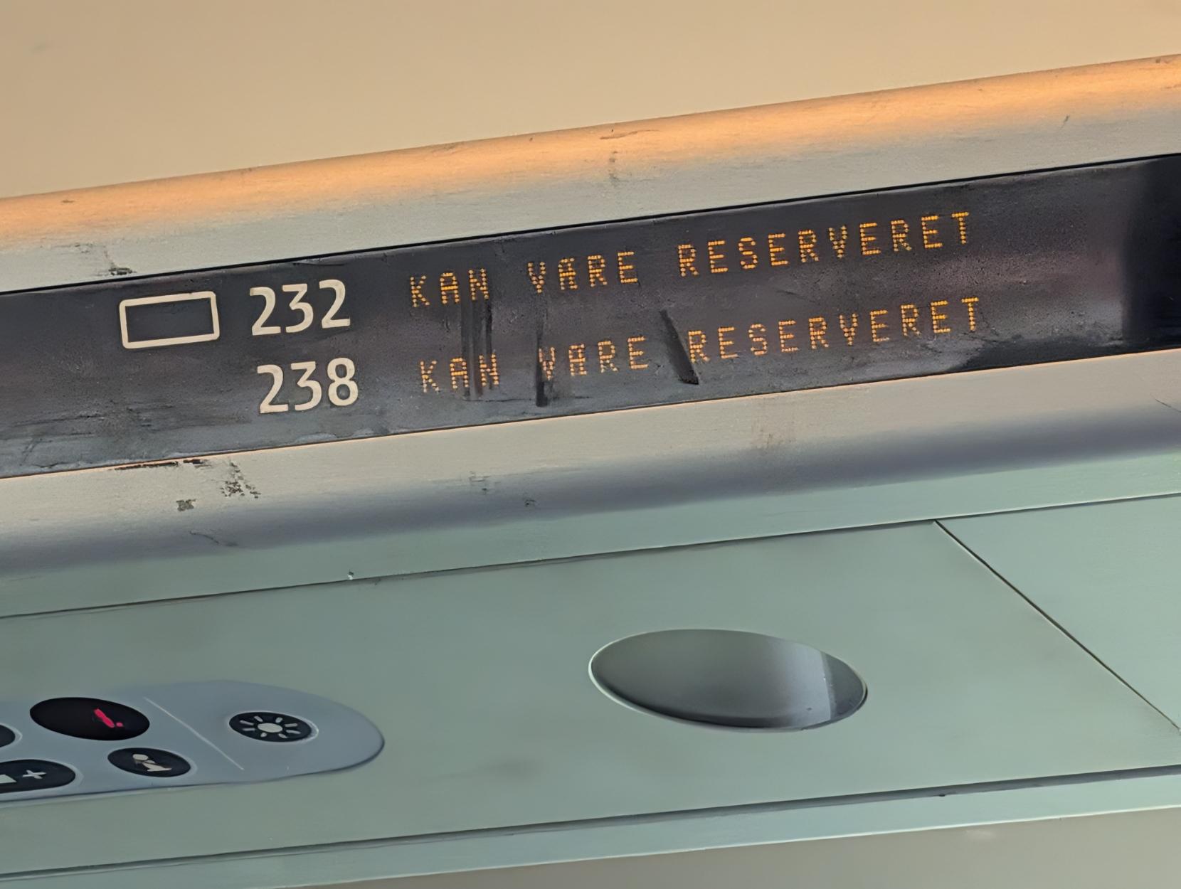 Eit nærbilete av eit digitalt setereservasjonsskilt inne i kupéen på eit dansk DSB IC4-tog. Skiltet er integrert i bagasjehylla over seta og viser to setenummer i kvit skrift: «232» og «238». Ved sidan av kvart nummer lyser ein oransje punktmatrisetekst med den danske meldinga «KAN VÆRE RESERVERET». Under skiltet kan ein sjå ein del av det lysegrå panelet med lufteventilar og knappar for leselys.
