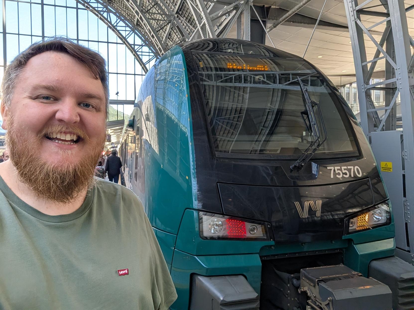 Ein selfie av ein breitt smilande mann med skjegg og salviegrøn t-skjorte inne på Bergen stasjon. Til høgre står den mørkegrøne og svarte fronten på eit elektrisk Vy-tog av typen Stadler FLIRT (Vy litra BM75 / Vossebanen-sett). Togfronten har Vy sin logo i sølv, og over det venstre markeringslyset (som lyser raudt) står settnummeret «75570». Det digitale destinasjonsskiltet øvst i frontruta viser teksten «Ikke i trafikk». I bakgrunnen reiser den store bogehalden i stål og glas seg over perrongen, der ein kan sjå ein person som går i bakgrunnen.