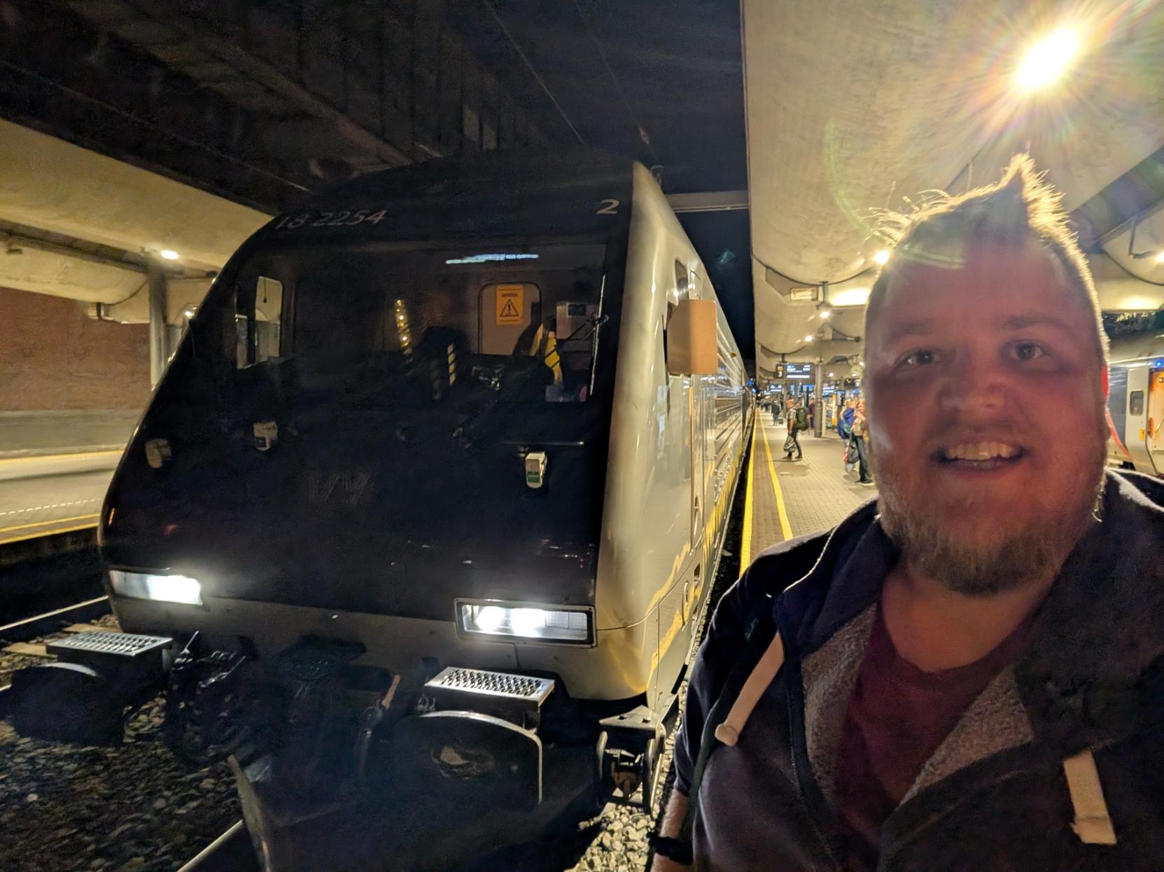 Ein selfie av ein breitt smilande mann med skjegg, mørkeblå hettejakke med lodden krage og vinraud t-skjorte, med skulderreimene på ryggsekken synlege, på ein overbygd perrong på Oslo Sentralstasjon (Oslo S) om kvelden. Til venstre står fronten på eit mørkegrått elektrisk lokomotiv av typen El 18 (SLM/Adtranz), som skal dra toget over Bergensbanen til Bergen. Lokomotivet har det karakteristiske, hellande frontpartiet og er merka med loknummeret «18 2254» i kvit skrift øvst over frontruta, og eit «2» ute på hjørnet. Frontlysa lyser skarpt, og i førarrommet skimtar ein instrumentpanel og eit gult og svart varselskilt. Bak lokomotivet står dei kvite og grå vognene med gul stripe langs sida, dekte av ein fotodekor med teksten «SE NORGE. TA TOGET». I bakgrunnen går den gule tryggleikslinja bortover perrongen mot folk, avgangstavler og eit anna togsett, under eit lågt betongtak der ei lampe blenkjer bak hovudet hans.

🚆 Tog: El 18 · Nr: 18 2254 · Stasjon: Oslo S · Bane: Bergensbanen · Selskap: Vy
