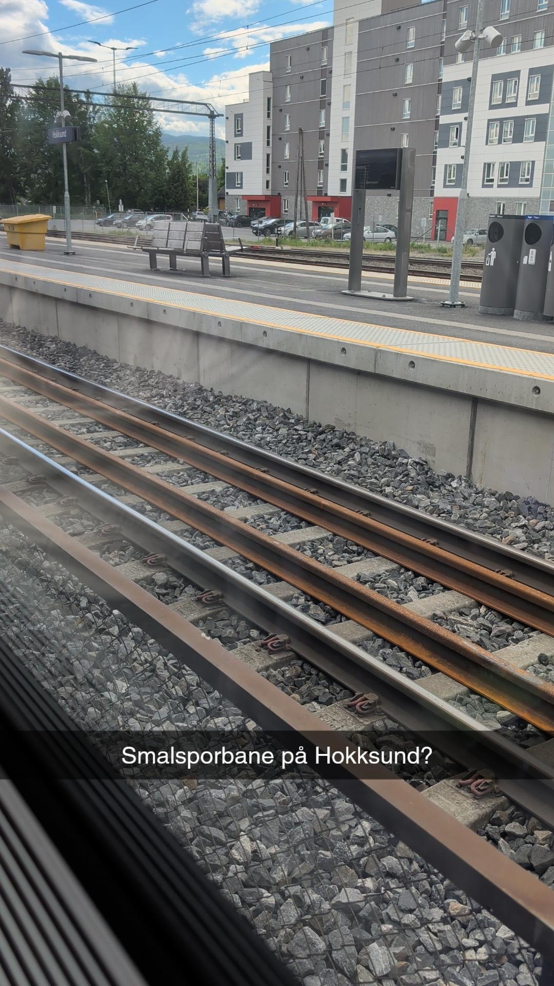 Eit bilete teke innanfrå eit togvindauge som viser jernbanesporet og perrongen på Hokksund stasjon. I føregrunnen ser ein togskjenene og pukksteinen i sporet, i tillegg til eit ekstra sett med skjenene som ligg tett inntil kvarandre. Midt på biletet er det lagt inn ein mørk tekstboks med kvit skrift der det står «Smalsporbane på Hokksund?». På den folketomme perrongen i bakgrunnen står ein benk, søppelkasser og ein rutetabellskjerm. Bak stasjonen ligg det moderne, grå og kvite blokker med raude detaljar under ein delvis blå himmel med kvite skyer.