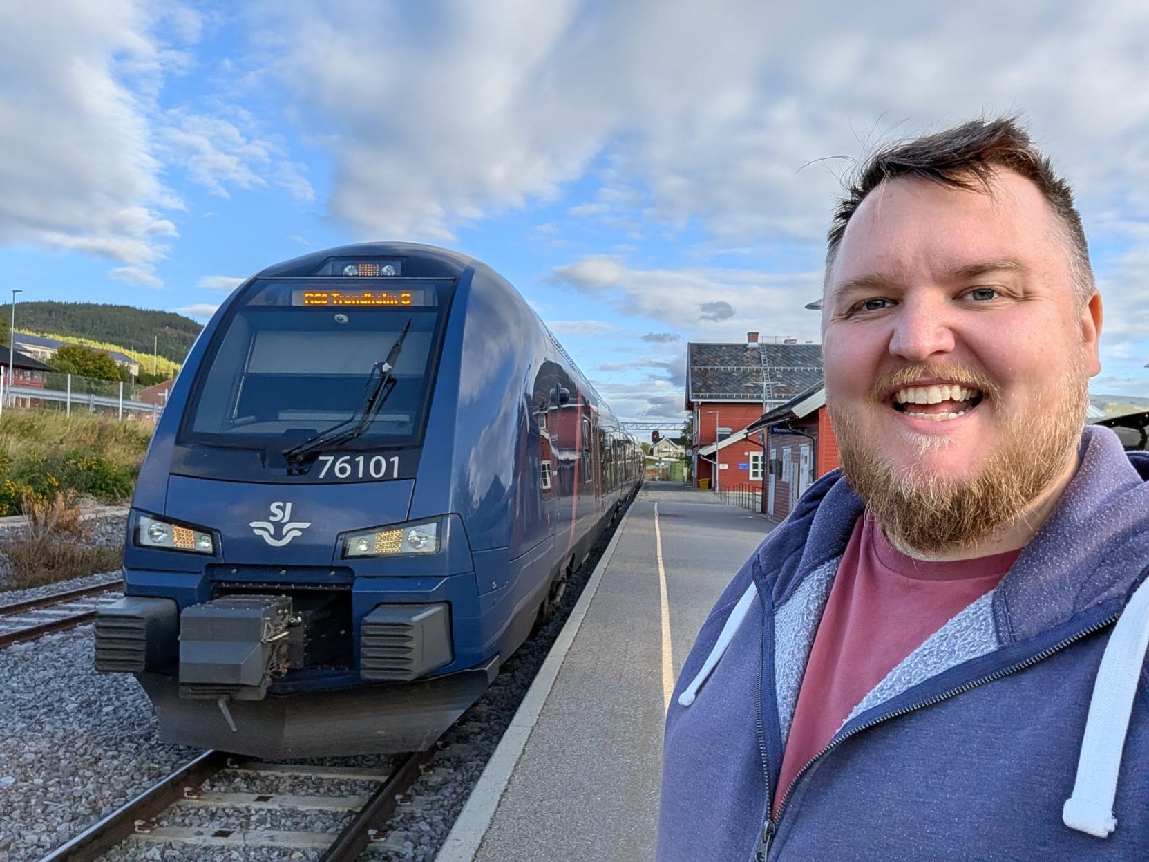 Ein selfie av ein breitt smilande mann med skjegg, raudrosa t-skjorte og lilla hettejakke med ljos fôrkant på perrongen på Alvdal stasjon. Til venstre står fronten på eit mørkeblått bimodalt motorvognsett av typen Stadler FLIRT (SJ Nord litra BM76), klart til avgang med R60 mot Trondheim. Over frontruta lyser destinasjonsskjermen med teksten «R60 Trondheim S» i oransje, settnummeret «76101» står i kvit skrift under ruta, og SJ-logoen med det vinga symbolet sit midt på frontpartiet. Frontlysa og dei gule markeringslysa lyser, og ei raud stripe følgjer vognsida bakover. I bakgrunnen til høgre ligg den raudmåla, gamle stasjonsbygningen med skiferttak, og til venstre reiser ein skogkledd ås seg mot ein blå kveldshimmel med kvite skyer.

🚆 Tog: Type 76 · Litra: BM76 · Nr: 76101 · Stasjon: Alvdal · Bane: Rørosbanen · Selskap: SJ Nord