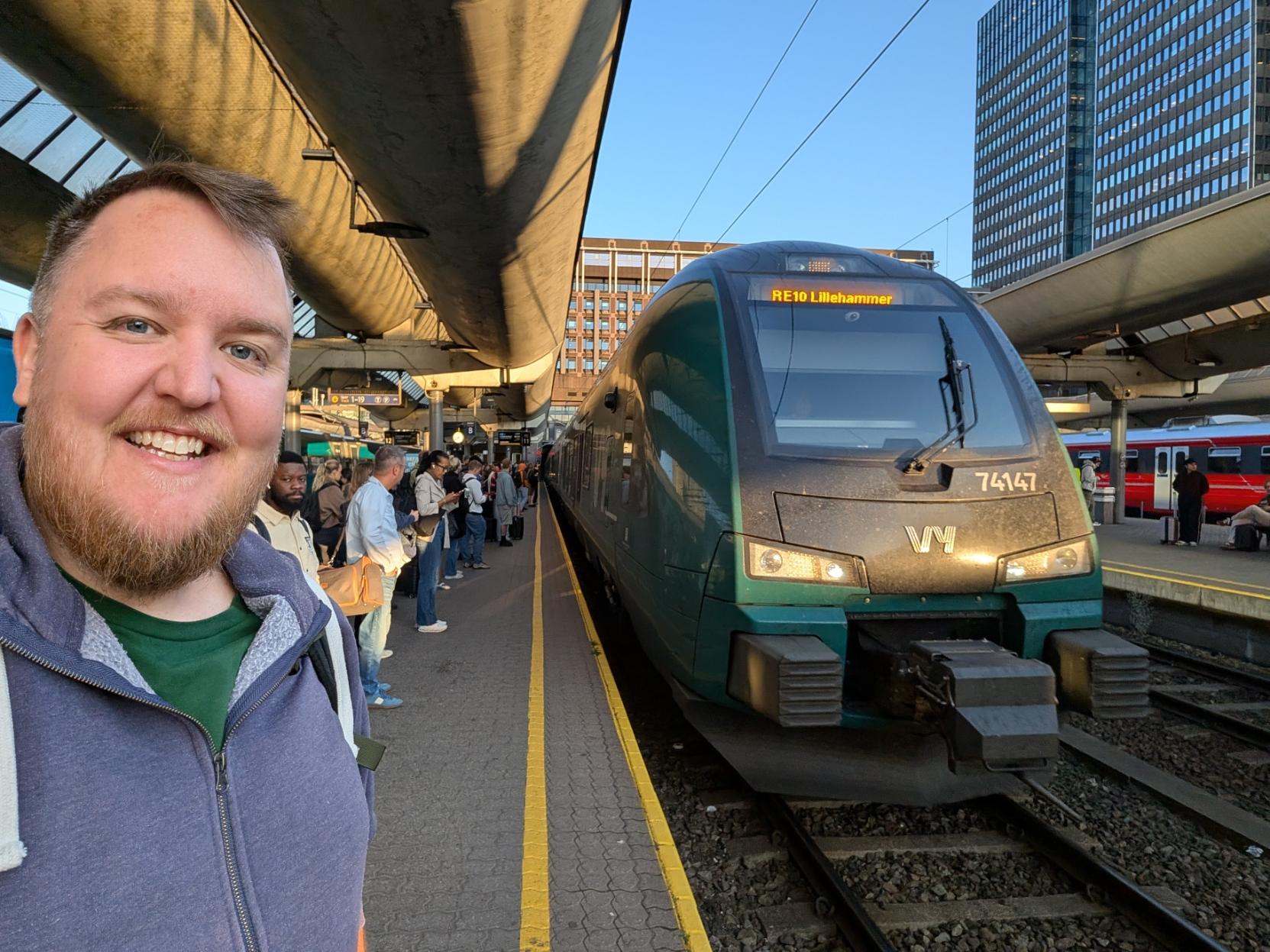 Ein selfie av ein breitt smilande mann med skjegg, grøn t-skjorte og gråblå hettejakke på ein overbygd perrong på Oslo Sentralstasjon (Oslo S). Til høgre står fronten på eit grønt og mørkegrått elektrisk Vy-tog av typen Stadler FLIRT (Vy litra BM74), merka med settnummeret «74147» under frontruta og Vy sin kvite logo på snuten. På destinasjonsskjermen over frontruta står det «RE10 Lillehammer», og frontlysa lyser mot ein støvete front. Den gule tryggleikslinja går langs perrongkanten. I bakgrunnen ventar ei rekkje folk på perrongen, eit raudt togsett står ved nabospora, og kontorbygga i Oslo sentrum reiser seg mot ein klår, blå kveldshimmel.

🚆 Tog: Stadler FLIRT · Litra: BM74 · Nr: 74147 · Stasjon: Oslo S · Bane: RE10 · Selskap: Vy