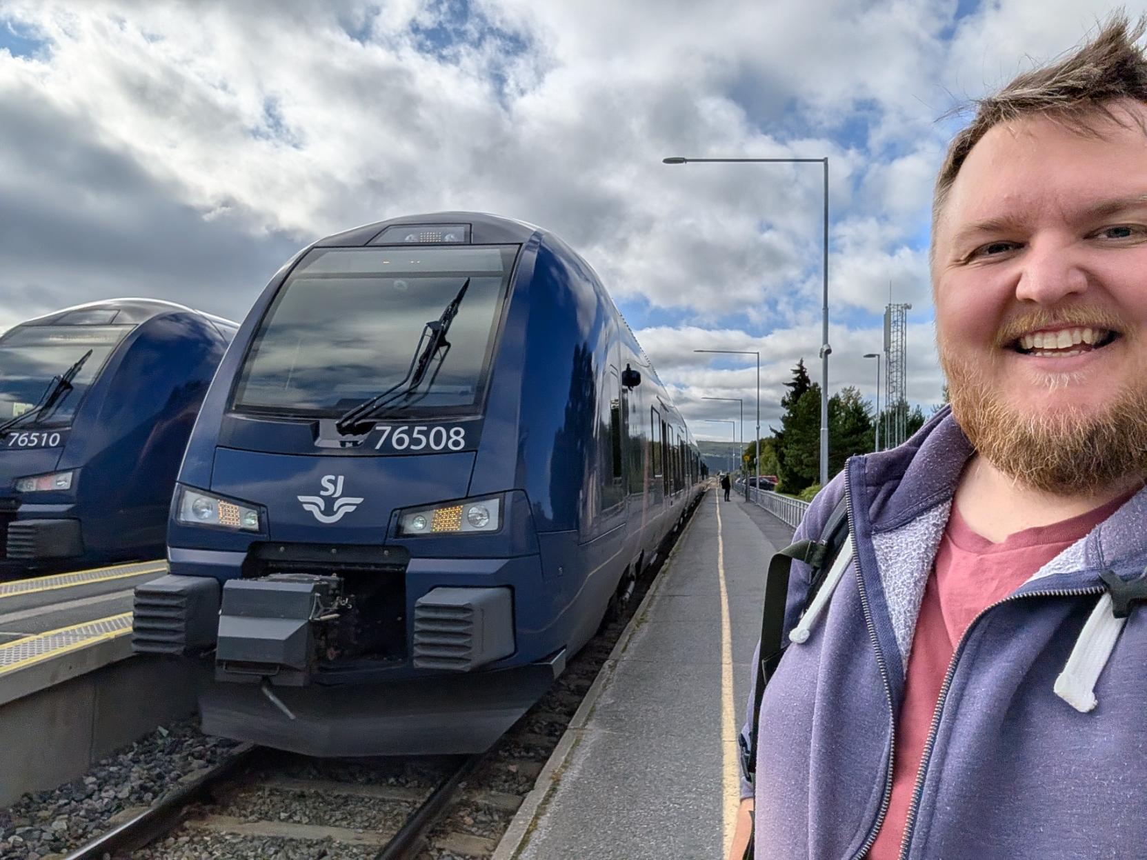 Ein selfie av ein breitt smilande mann med skjegg, lilla hettejakke og vinraud t-skjorte, med ei ryggsekkreim over skuldra, på perrongen på Røros stasjon. Til venstre står fronten på eit mørkeblått bimodalt motorvognsett av typen Stadler FLIRT (SJ Nord litra BM76), merka med settnummeret «76508» under frontruta og SJ-logoen med det vinga hjulet midt på snuten. Frontlysa og dei oransje markeringslysa lyser, og settet står klart til å køyra R60 over Rørosbanen mot Hamar. På nabosporet bortanfor står eit søstersett merka «76510». Den gule tryggleikslinja følgjer den nesten tomme perrongen bakover forbi ei rekkje lyktestolpar, ein telemast og granskog, under ein blå himmel med store, kvite skyer.

🚆 Tog: Stadler FLIRT (Type 76) · Litra: BM76 · Nr: 76508 · Stasjon: Røros · Bane: Rørosbanen (R60) · Selskap: SJ Nord