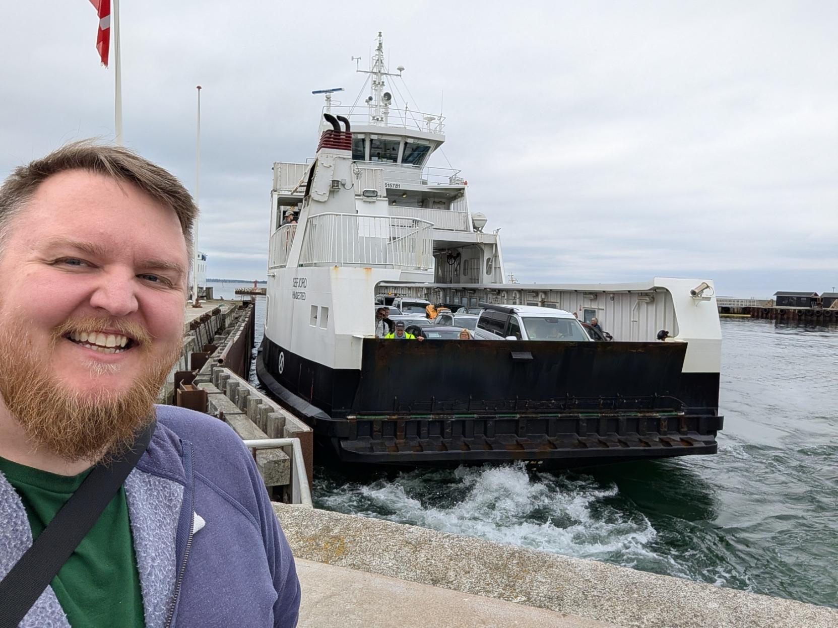Ein selfie av ein breitt smilande mann med skjegg og lilla hettegenser som står på kaia. Rett bak han klappar den kvite og svarte bilferja M/F Isefjord til kai, og det kvite skummet i vatnet viser at ho rygger eller bremsar opp mot ferjeleiet. På bildekket kan ein sjå fleire parkerte bilar og passasjerar som ventar på å køyre i land. Himmelhvelvingen er heilt grå og overskya, og landskapet rundt ber preg av eit ope hamneområde.