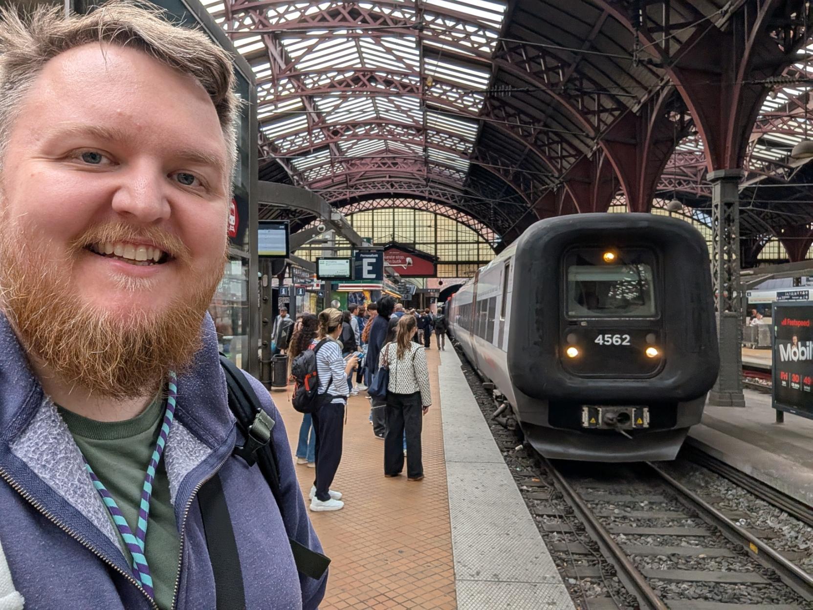 Ein selfie av ein breitt smilande mann med skjegg og lilla hettegenser på ein travel perrong inne på Københavns Hovedbanegård. Til høgre står fronten på eit grått og kvitt elektrisk Øresundstog (litra ET) på sporet. Togfronten har det karakteristiske designet med ein tjukk, svart gummimembran rundt frontruta, og er merka med tognummeret «4562». Langs perrongen til venstre står det ein stor flokk med reisande som ventar på å gå om bord. I bakgrunnen breier den imponerende, historiske stasjonshallen i tre og stål seg ut over spora.