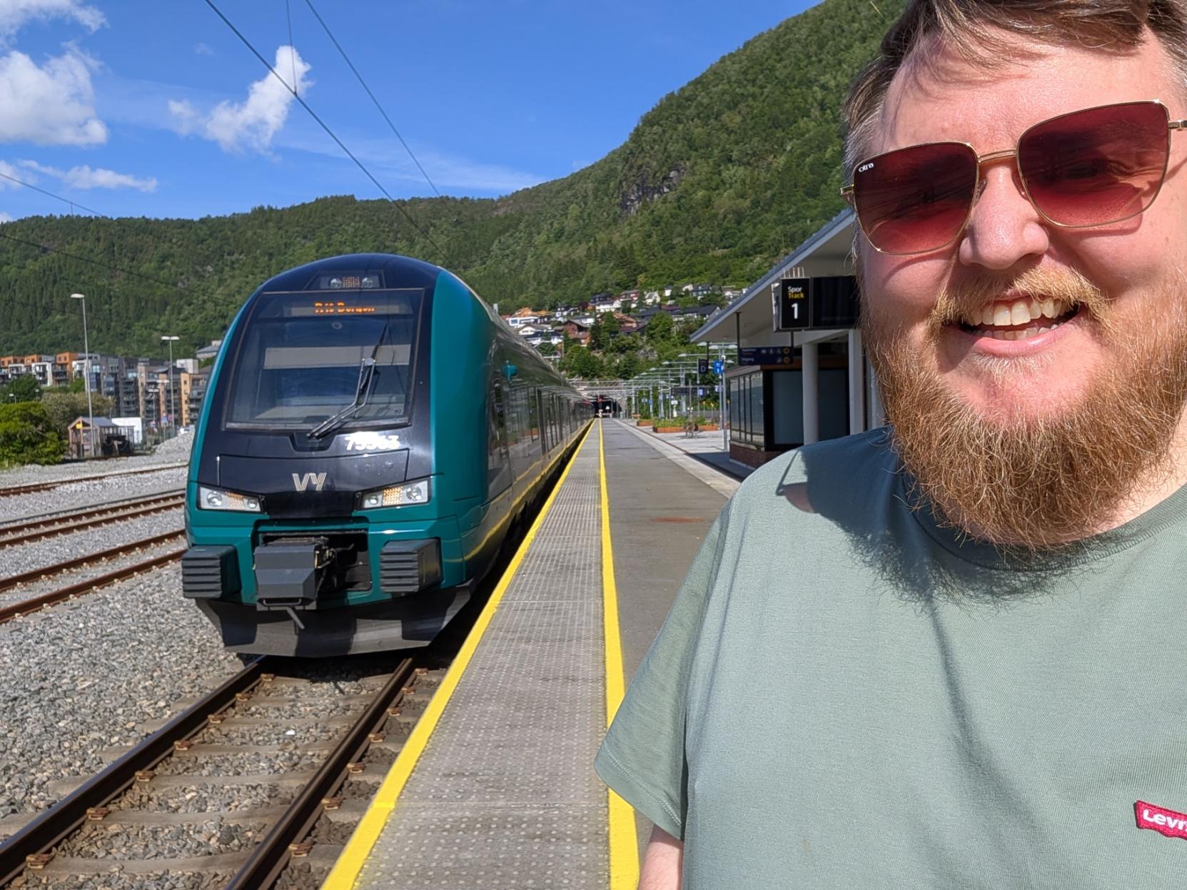 Ein selfie av ein smilande mann med skjegg, solbriller og ein salviegrøn t-skjorte på ein solfylt perrong på Arna stasjon (Spor 1). Til venstre står fronten på eit grønt og mørkegrått elektrisk Vy-tog av typen Stadler FLIRT (Vy litra BM75) klart ved plattforma, merka med settnummeret «75563» under frontruta. Den gule tryggleikslinja strekkjer seg langs den tomme perrongen ved sidan av toget. I bakgrunnen ser ein bustadhus i åssida og eit grønt, skogkledd fjell som reiser seg mot ein klår, blå sommarhimmel med nokre få, kvite skyer.