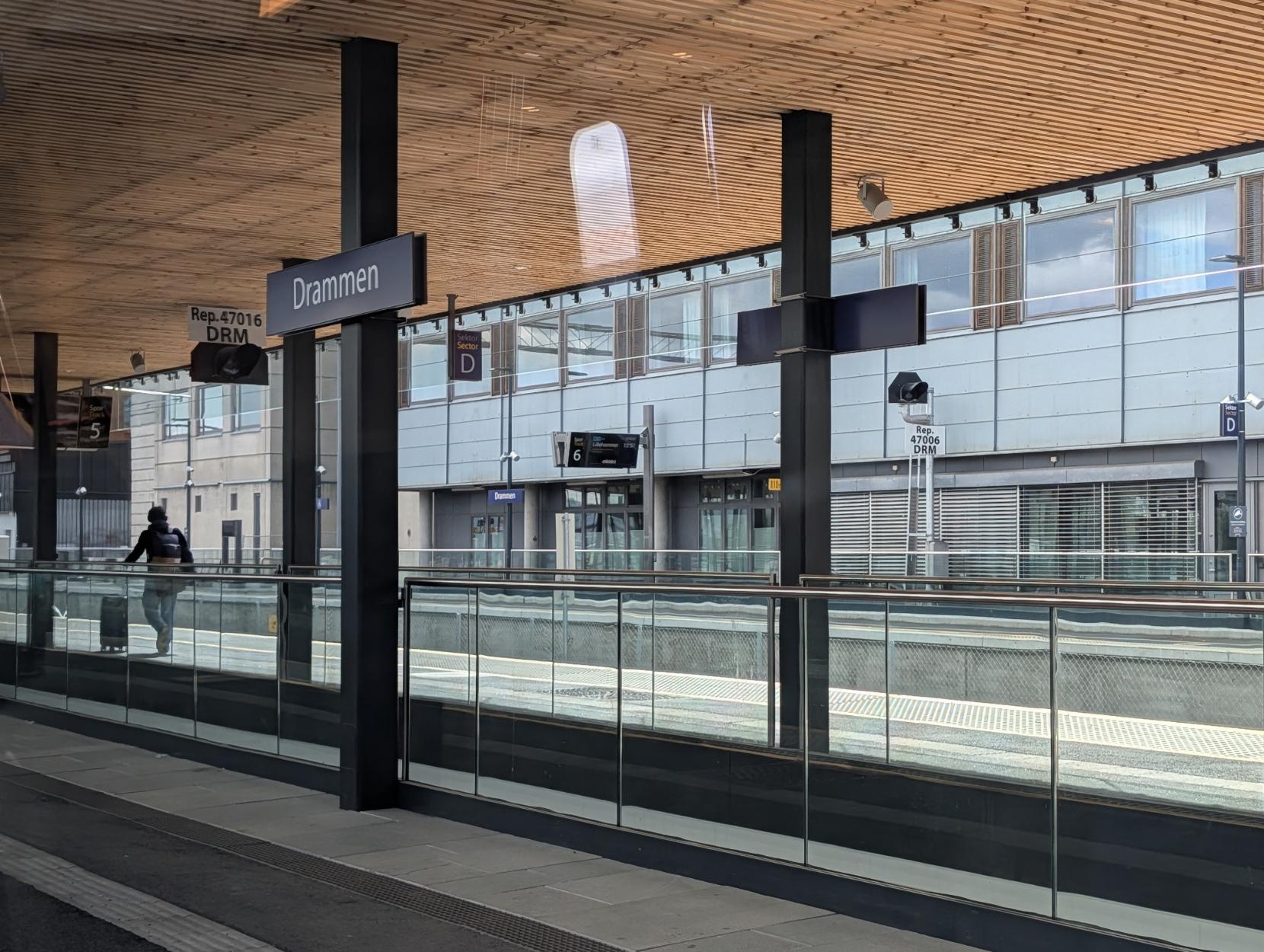 Eit bilete teke frå ein overbygd perrong på Drammen stasjon. I føregrunnen ser ein eit rekkverk i glas og metall som skil perrongane, og eit skilt på ein berande stolpe der det står «Drammen». Til venstre går ein person med ryggsekk bortover perrongen. Stasjonstaket har ein lys, moderne trepaneloverflate som speglar seg i glasflatene. I bakgrunnen ser ein spora, digitale informasjonsskilt (mellom anna for spor 6 med avgang mot Dal), og den grå fasaden til stasjonsbygningen med store vindaugo.