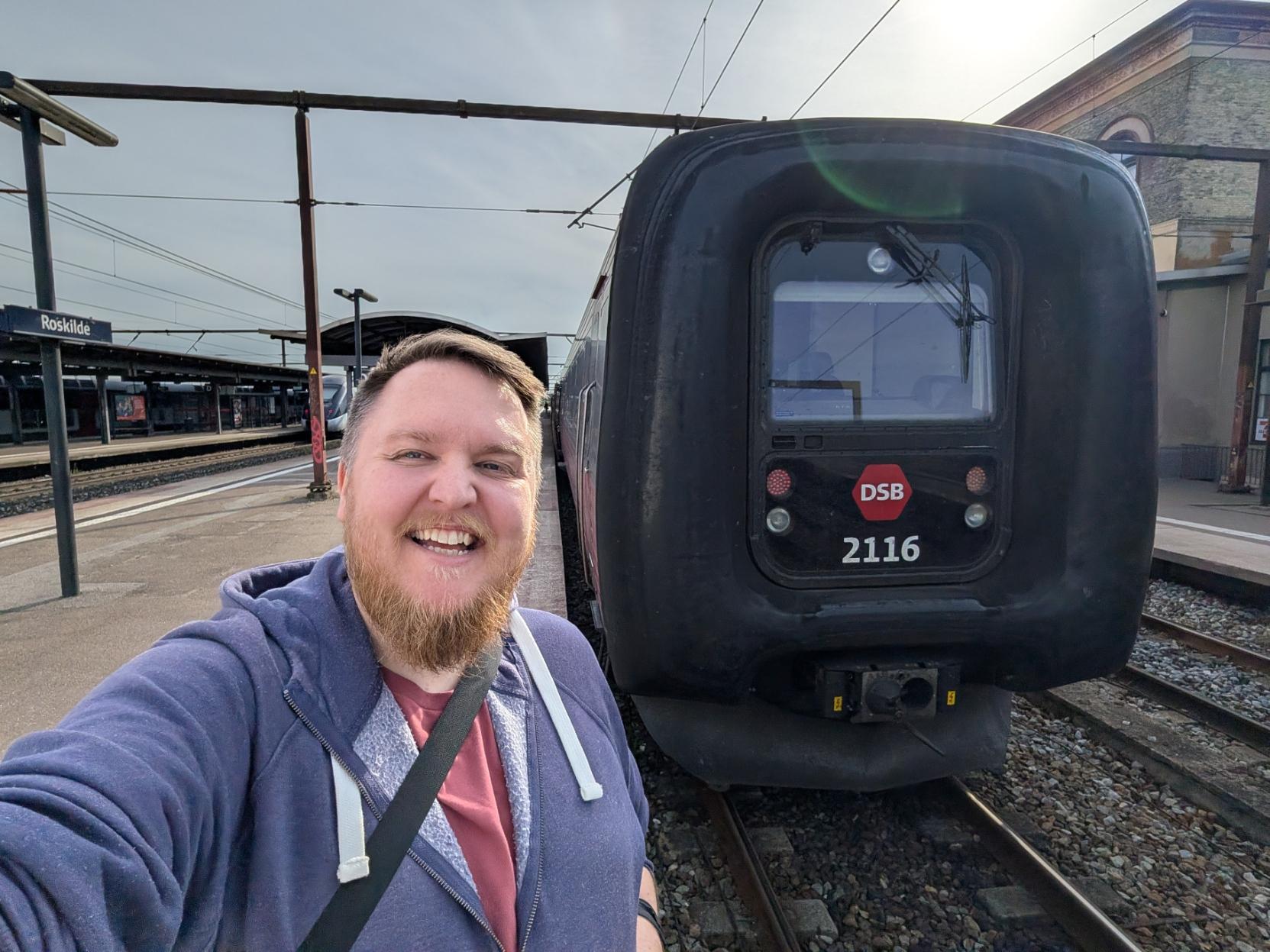 Ein selfie av ein breitt smilande mann med skjegg og lilla hettegenser på ein perrong på Roskilde stasjon i solskinn. Til høgre står den karakteristiske fronten på eit DSB IC3-tog på sporet. Togfronten er prega av den store, svarte gummimembranen som omkransar førarromsvindauget, og er merka med DSB sin raude logo og tognummeret «2116». I bakgrunnen ser ein stasjonsarkitekturen med perrongtak, eit anna tog lenger unna til venstre, stasjonsskiltet der det står «Roskilde», og ein eldre stasjonsbygning i mur til høgre under ein ljos himmel.