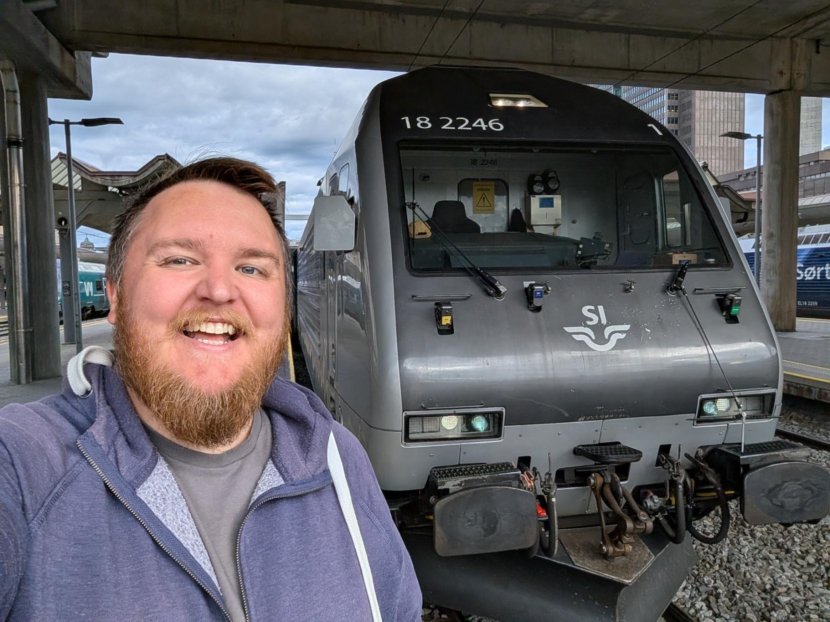 Ein selfie av ein breitt smilande mann med skjegg og blå hettejakke på ein perrong under tak på Oslo Sentralstasjon (Oslo S). Til høgre står fronten på eit mørkegrått elektrisk lokomotiv av typen El 18 (SNCL/SLM/Adtranz), som dreg F6-toget på Dovrebanen mot Trondheim. Lokomotivfronten har det karakteristiske, hellande designet og er merka med lokomotivnummeret «18 2246» øverst over frontruta. Midt på frontpartiet prydar den kvite SJ Nord-logoen. Frontlysa på lokomotivet lyser, og i førerrommet skimtar ein eit gult og svart varselskilt samt instrumentpanel. I bakgrunnen til venstre kan ein sjå andre togsett på perrongane, og over perrongtaket hevar næringsbygga i Oslo sentrum seg mot ein delvis overskya himmel.