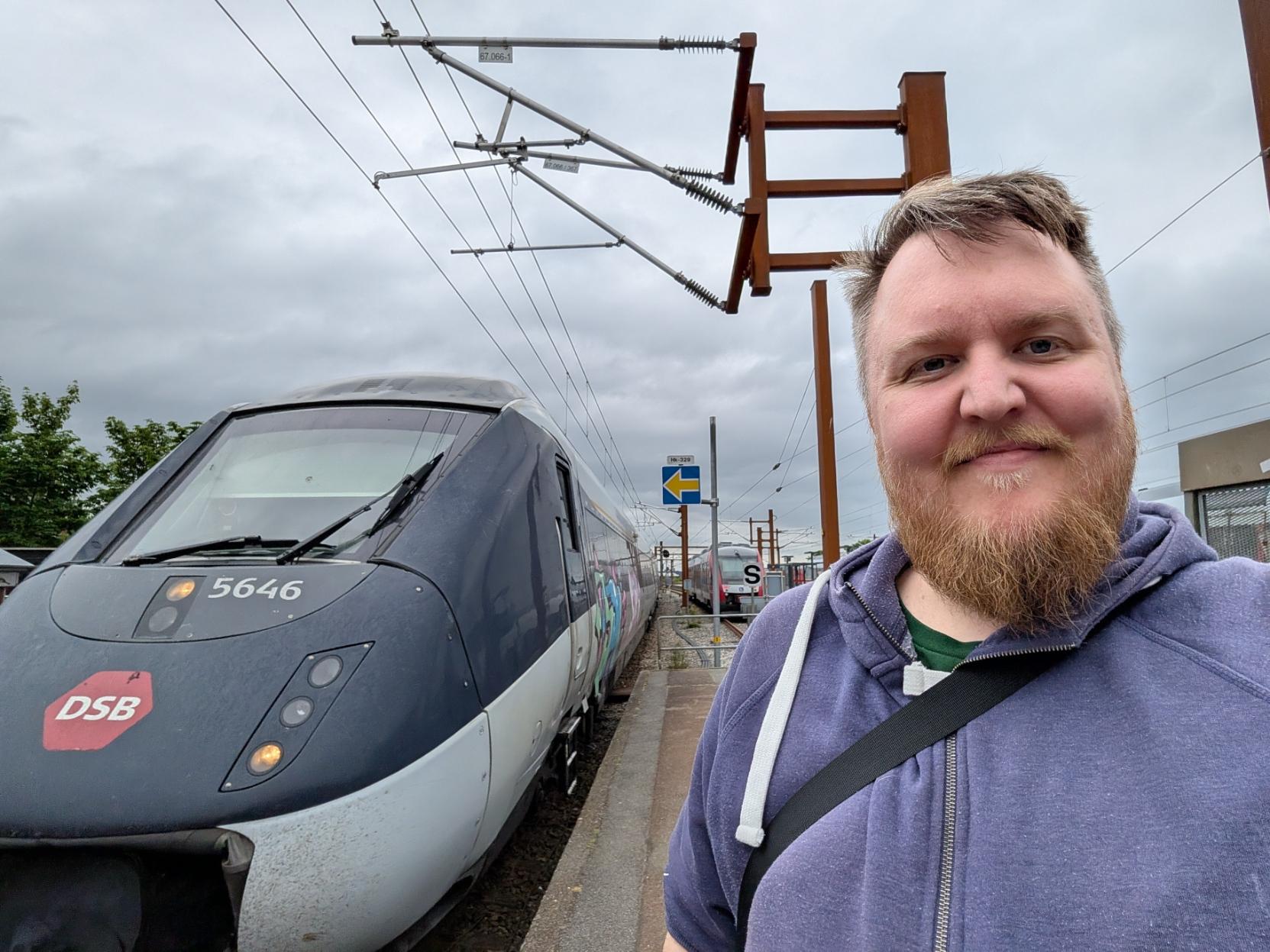 Ein selfie av ein smilande mann med skjegg og lilla hettegenser på ein utandørs perrong. Til venstre står den spisse, mørkeblå og grå fronten på eit DSB-tog av modellen IC4 (klasse MG), med tognummeret 5646 og DSB sin raude logo tydeleg synleg på snuten. Sida på togsettet har noko fargerik grafitti. Over sporet heng det koplingsledningar til strømnettet, og i bakgrunnen anar ein eit anna tog, eit gult pilsymbolskilt, samt ein heilt grå og overskya himmel.