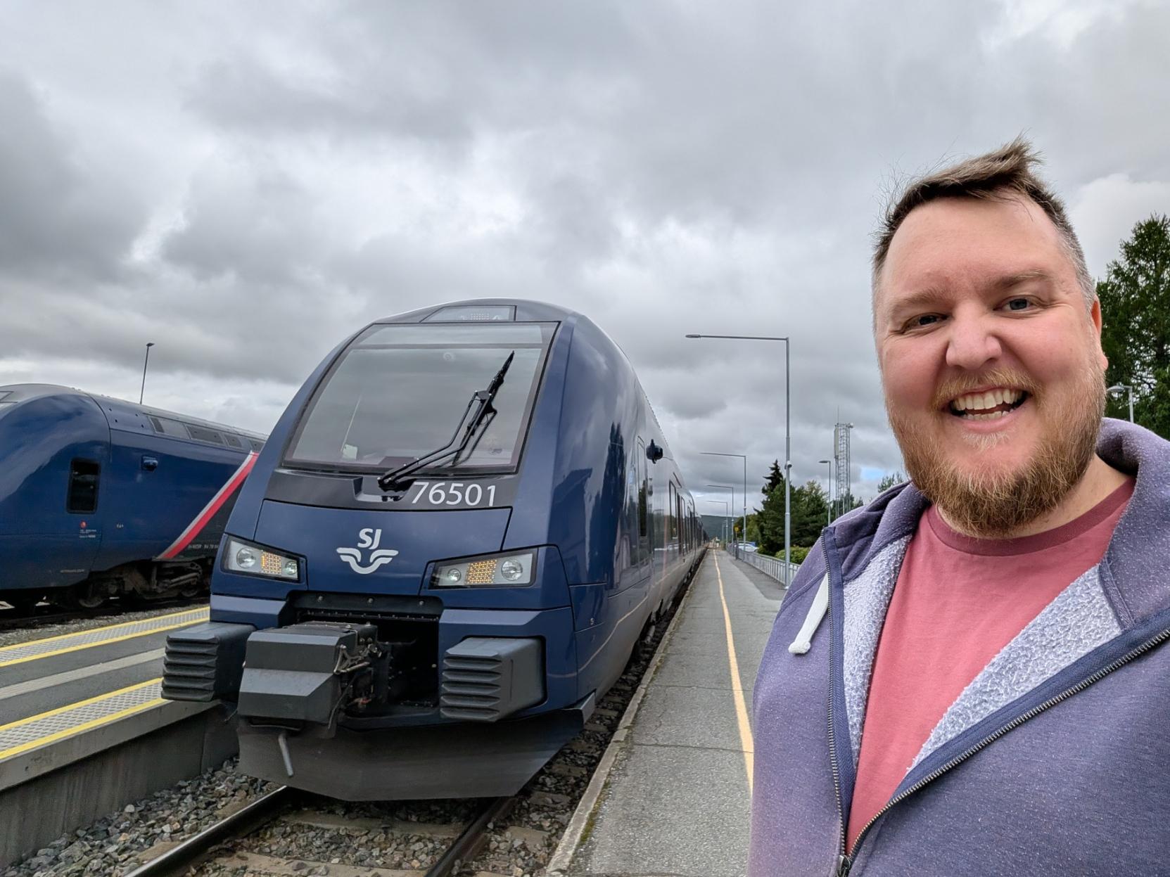 Ein selfie av ein breitt smilande mann med skjegg, raudrosa t-skjorte og lilla hettejakke med ljos fôrkant på perrongen på Røros stasjon. Til venstre står den avrunda fronten på eit mørkeblått bimodalt motorvognsett av typen Stadler FLIRT (SJ Nord litra BM76), merka med settnummeret «76501» under frontruta og SJ-logoen med det vinga symbolet midt på frontpartiet. Frontlysa og dei gule markeringslysa lyser, og den gule tryggleikslinja følgjer perrongen bortover langs toget. På nabosporet heilt til venstre står fronten på eit anna sett av same typen. I bakgrunnen ser ein perronglysstolpar, grøne tre og ei mast, under ein tung, grå og overskya himmel.

🚆 Tog: Type 76 · Litra: BM76 · Nr: 76501 · Stasjon: Røros · Bane: Rørosbanen · Selskap: SJ Nord
