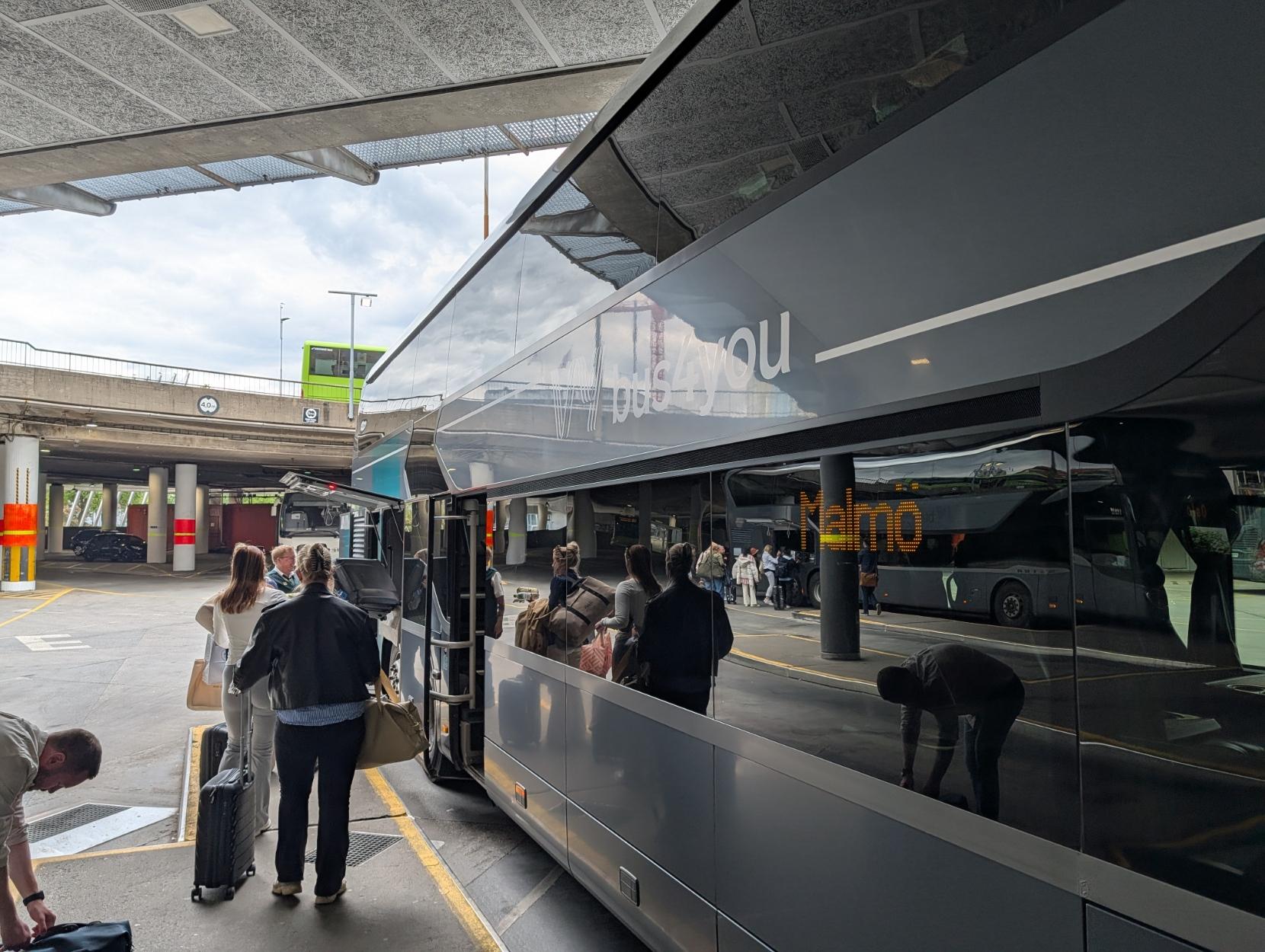 Ein mørkegrå Setra S 531 DT dobbeltdekkarbuss står parkert på ein overbygd bussterminal, der passasjerar står i kø for å gå om bord. På bussen sin blanke sidevegg står logoen «Vy bus4you» i kvit skrift. Dei store vindaugo speglar terminalen, dei reisande og eit gult digitalt skilt der det står «Malmö». Til venstre i biletet står fleire personar med bagasje, og ein svart trillekoffert er synleg i føregrunnen. I bakgrunnen ser ein betongkonstruksjonar og ein annan grøn buss på eit øvre nivå.