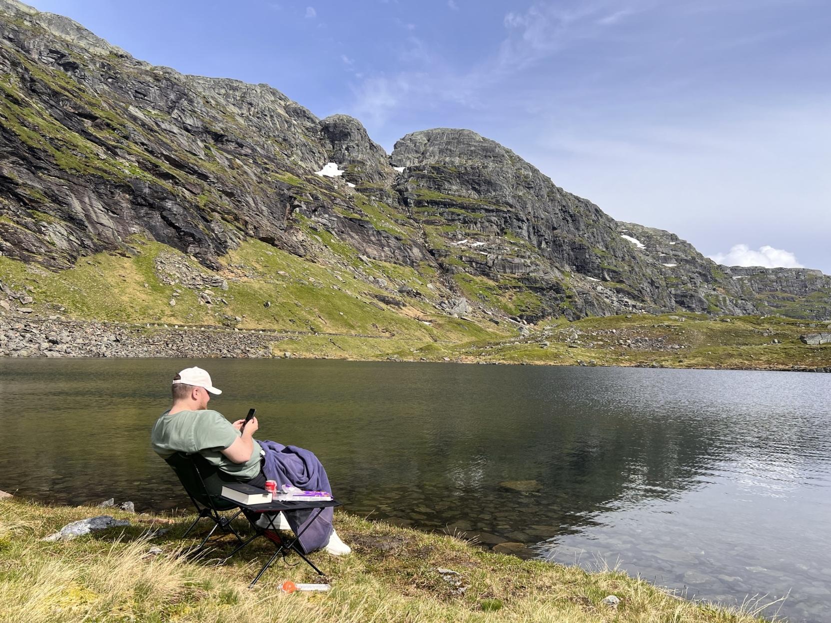 Eit utendørsbilete i klårt dagslys som viser ein mann sett frå sida der han sit i ein campingstol på den grasdekte breidda av Elversvatnet i Røldal. Mannen har på seg ein salviegrøn t-skjorte og ein ljos caps, sit tilbakelent med eit lilla teppe (faste lesare kjenner det att som ein hettegenser) over beina, og ser ned på ein mobiltelefon han held i hendene. Framfor stolen står eit lite, samanleggbart campingbord der det ligg ei bok, ein pakke kjeks, og ein boks cola. Det blikstille fjellvatnet speglar dei mektige, bratte fjellsidene i bakgrunnen, som er prega av grå stein, grøne grassletter og enkelte små snøflekkar under ein klår, ljos blå himmel.