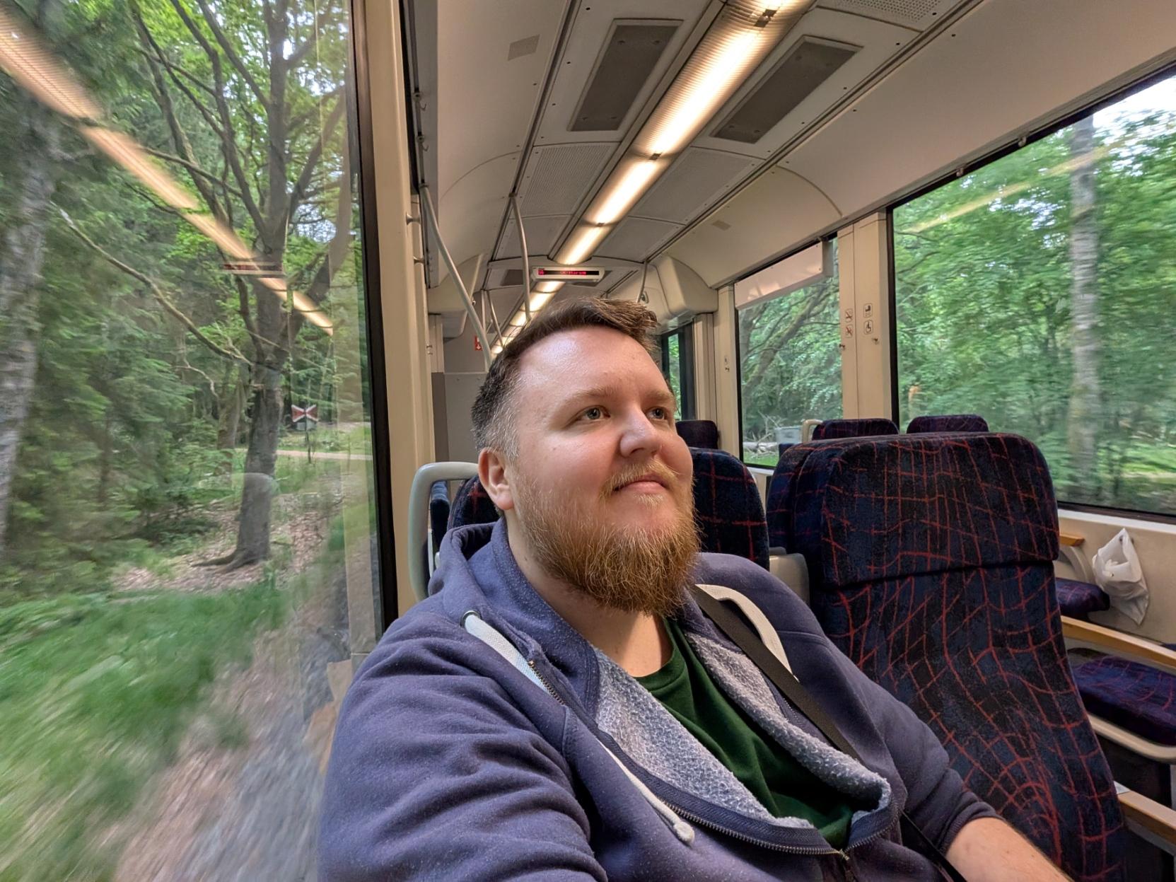 Ein selfie av ein mann med raudbrunt skjegg og blå hettegenser som sit i eit passasjersete inne på eit lokaltog av modellen Alstom Coradia LINT 41. Mannen smiler svakt og kikkar opp og framover i kupéen. Toget har mørkeblå passasjersete med eit raudt og lyst mønster. Det er store panorama-vindaugo på begge sider av vogna, og på utsida fyk ein tett, grøn og frodig skog forbi i full fart, noko som gir ein tydeleg fartsfølelse i biletet. Kupéen er lys og open med langstrakte lysrøyr i taket.