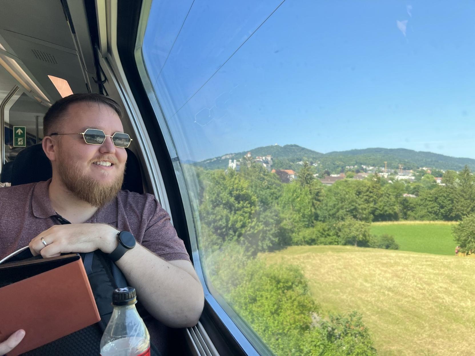 Bilete av mann (@markus@skvip.lol) i ei togvogn. Han har på solbrillar og ser ut på eit grønt landskap med bygg på berga. Blå himmel.