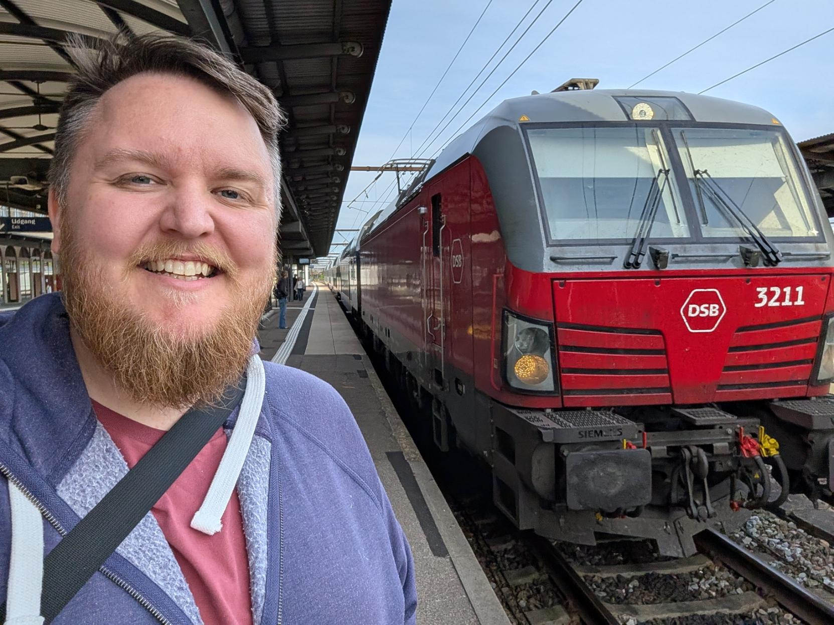Ein selfie av ein breitt smilande mann med skjegg og lilla hettegenser på ein overbygd togperrong. Til høgre står fronten på eit raudt og grått elektrisk lokomotiv av typen Siemens Vectron (DSB litra EB) på sporet. Lokomotivet er merka med DSB sin kvite logo på den raude fronten, har lokomotivnummeret «3211» oppe til høgre, og ein kan sjå «SIEMENS»-preginga på fofsplata. Lokomotivet er kopla til ei rekkje med passasjervogner som strekkjer seg bakover langs perrongen. I bakgrunnen ser ein perrongen fortsetje under taket, med nokre få personar i det fjerne, under ein ljos himmel