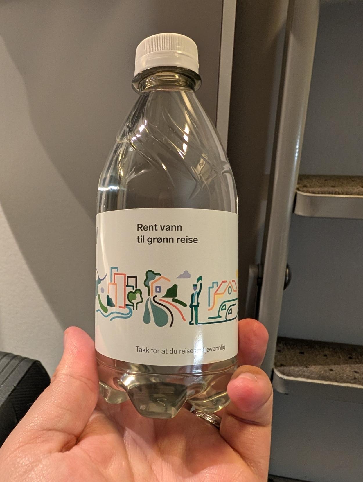Ein person held ei gjennomsiktig vassflaske av plast i handa. Flaskelappen har ei fargerik, enkel teikning av bygningssilingar, tre, vegar og ein togkonduktør. På lappen står teksten «Rent vann til grønn reise» øverst, og nederst står det «Takk for at du reiser miljøvennlig».