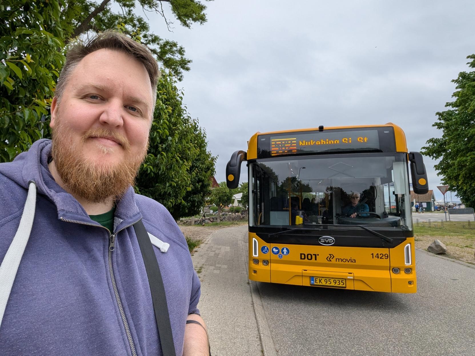 Ein selfie av ein smilande mann med raudbrunt skjegg og blå hettegenser som står ved sia av ein veg. Til høgre bak han står ein gul rutebuss av merket BYD (modell K9UB). Bussen er merka med DOT- og Movia-logoar samt vognnummeret 1429, og det digitale destinasjonsskiltet over frontruta lyser oransje med teksten «562 Nykøbing Sj St». Ein sjåfør er synleg bak rattet. Til venstre i biletet veks det grøne, tette tre, og i bakgrunnen ser ein ein asfaltert veg, nokre bygningar i det fjerne og ein grå, overskya himmel.