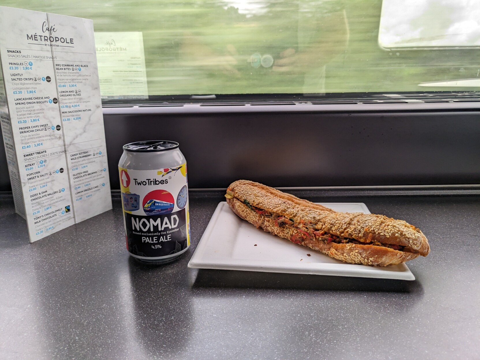 Bilete av ein meny, ein boks øl, og ein papptallerken med ein baguette på ein benk. Det er eit vindauge i bakgrunnen.

Menyen er merka «Café Métropole». Fleire smårettar er lista opp. 

 Boksølen er merka «Two Tribes Nomad Pale Ale.» Den har ein illustasjon av Eurostar-toget på seg. «Brewed exclusively for Eurostar.»

Baguetten ser vassen ut. Masse frø på toppen, grønsakar tytar ut av sidane.

Ut vindauget er det masse grønt som farar forbi. Ein kan så vidt kimte @markus@skvip.lol sin refleksjon i vindauget.
