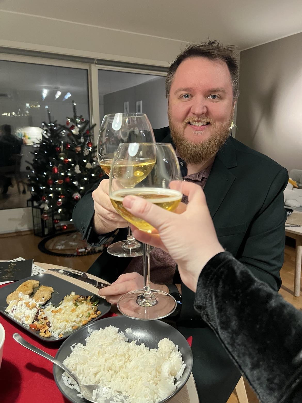 Bilete av Markus (@markus@skvip.lol) som skålar med eit vinglas med noko guleg (sprudlande te) i.

Markus har på seg raud skjorte og grøn blazer.

Det er eit pynta joletre i bakgrunnen.

Ein arm med svart kjoleerne skålar med Markus.

På middagsbordet mellom Markus og denne andre står ei skål med ris og ein tallerken med middag.