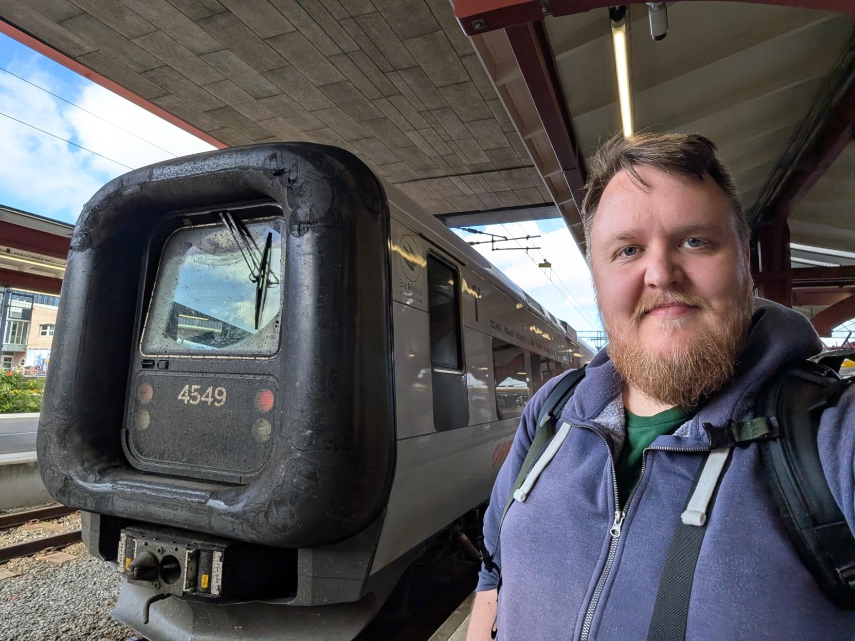Ein selfie av ein smilande mann med skjegg og blå hettegenser på ein overbygd perrong. Til venstre for han står fronten på eit grått Öresundståg av modellen X32K / X31K (Contessa) med nummeret 4549 tydeleg prega på snuten. Toget har sin karakteristiske, tjukke og avrunda gummifront (gummimansjett) som rammar inn frontruta og den framtidige overgangsdøra. I bakgrunnen ser ein stasjonsarkitektur, jernbanespor og ein delvis blå himmel over perrongtaket.