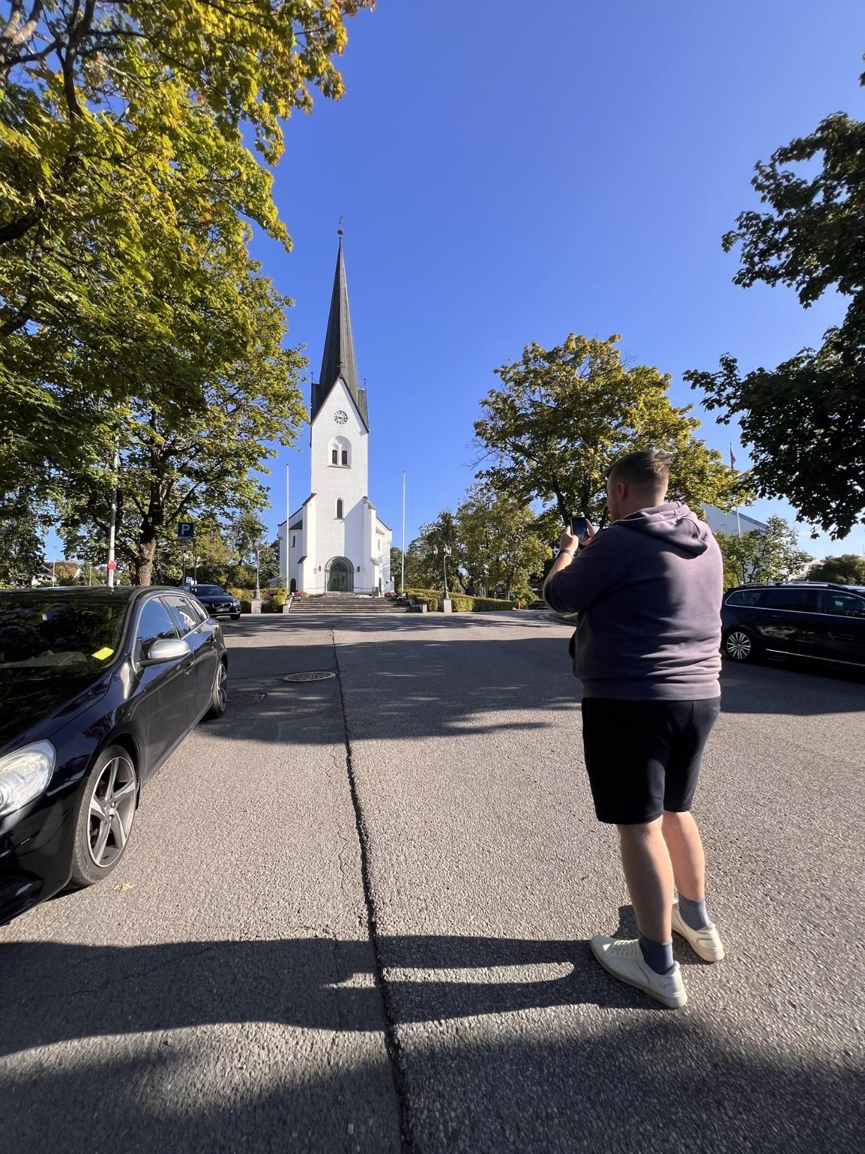 Ein mann i lilla hettegenser og shorts står på ein asfaltert veg og fotograferer ei kvit kyrkje med eit høgt, mørkt spir i enden av vegen. Det er solskin, og langs vegen står det grøne tre og parkerte bilar.