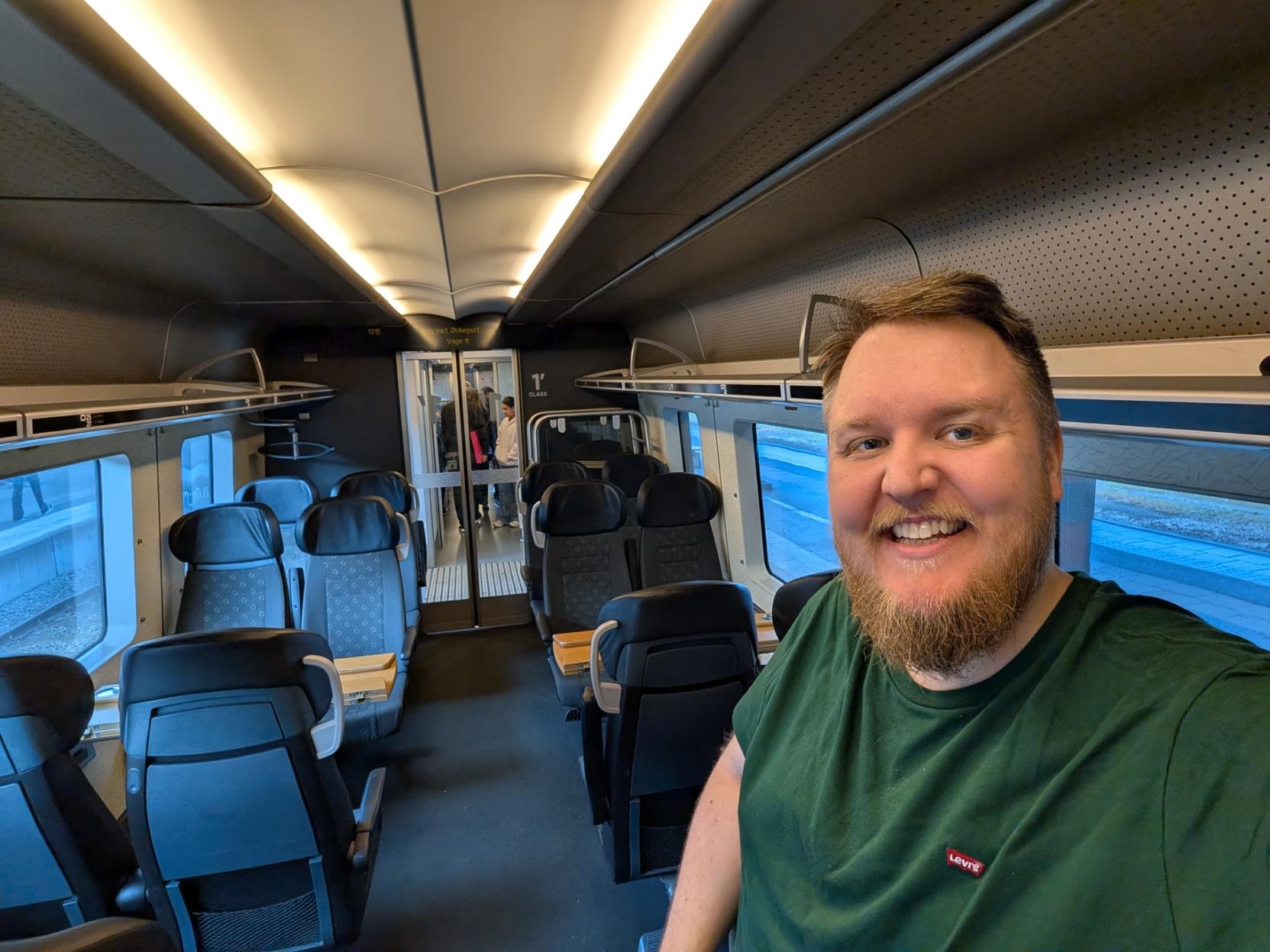 Ein selfie av ein smilande mann med skjegg og grøn t-skjorte inne i kupéen på eit Öresundståg av modellen X31K / X32K (Contessa). Mannen står til høgre i biletet, og bak han ser ein midtgangen og dei mørkegrå passasjerseta på 1. klasse (merka med «1st CLASS» på veggen). Seta er ordna to og to mot kvarandre med små trebord i mellom. Det er lyst og moderne inne i vognen med ein boga takbelysning. I bakgrunnen fører glasdører ut til ein plattformgang der andre passasjerar står, og gjennom togvindaugo på kvar side kan ein sjå glimt av perrongen utanfor.