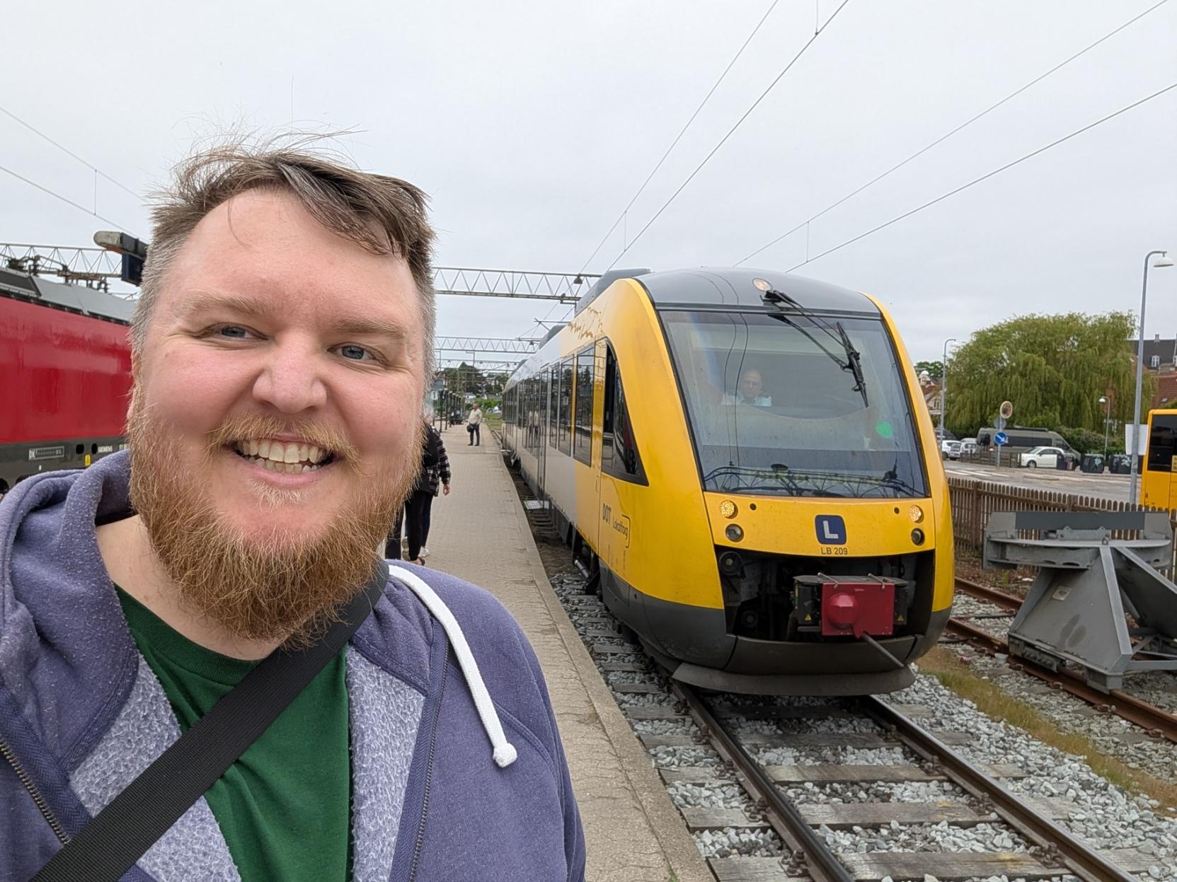 Ein selfie av ein breitt smilande mann med skjegg og blå hettegenser på ein utandørs perrong. Til høgre i biletet står fronten på eit gult og grått lokaltog av modellen Alstom Coradia LINT 41 (merka med ein blå «L»-logo og tognummer LB 209 på snuten). Ein lokførar er synleg gjennom den store frontruta. Til venstre anar ein kanten av eit anna raudt tog. Himmelhvelvingen er heilt overskya og kvit, og i bakgrunnen ser ein perrongen strekkje seg bakover med nokre få fotgjengarar, i tillegg til tre og master langs sporet.
