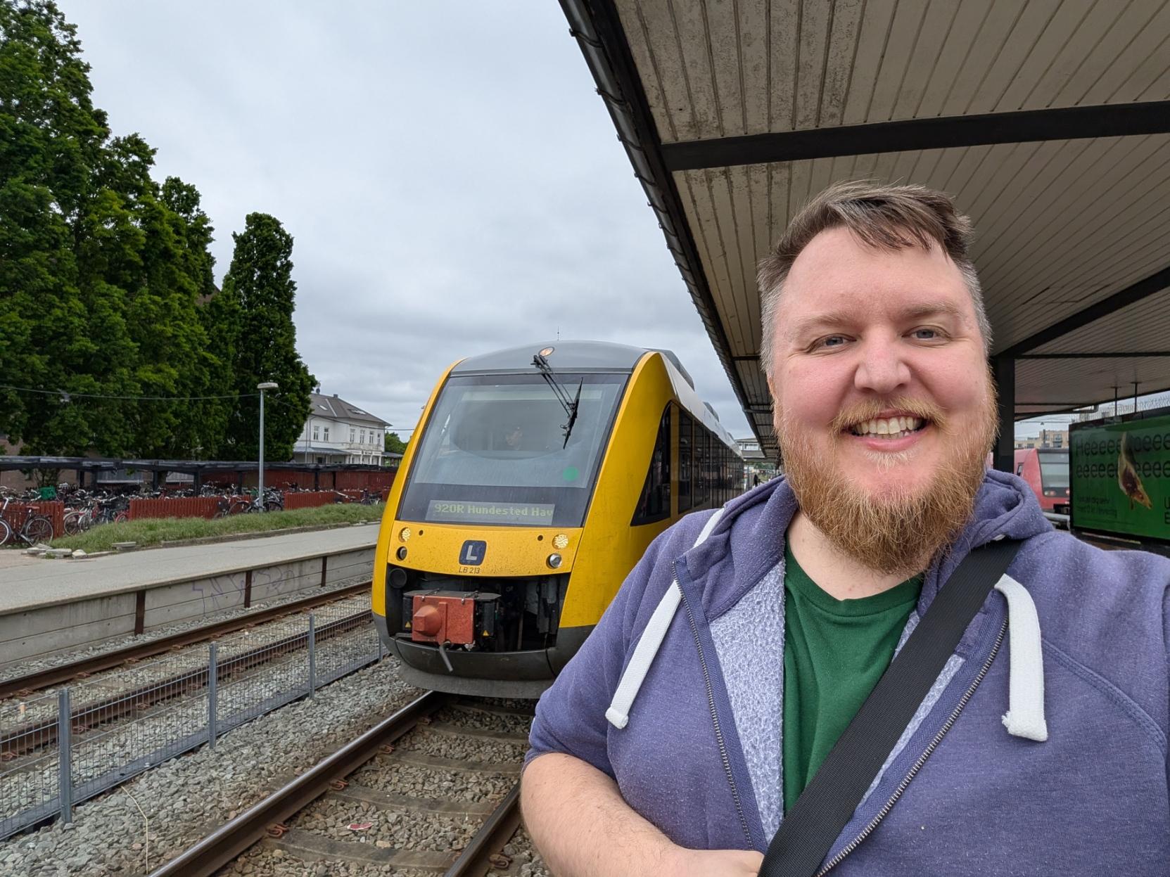 Ein selfie av ein breitt smilande mann med skjegg og lilla hettegenser på ein jernbanestasjon. Til venstre bak han står fronten på eit gult og grått lokaltog av modellen Alstom Coradia LINT 41 på sporet. Toget er merka med ein blå «L»-logo og har tognummeret LB 213 på snuten. Det digitale destinasjonsskiltet i frontruta viser teksten «920R Hundested Havn». Mannen står delvis under eit overbygd perrongtak til høgre. I bakgrunnen ser ein ein annan perrong med mange parkerte syklar, store grøne tre, ein kvit bygning og ein grå, overskya himmel.