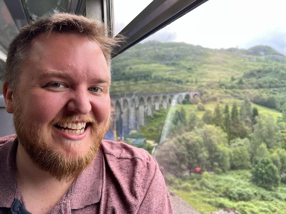 @markus@skvip.lol (kvit mann med mørkeblondt hår og skjegg) smilar ved togvindauget. I bakgrunnen er Glenfinnan-viadukten, kjent frå Harry Potter-filmane. 

Viadukten sprer seg over midten av biletet.

Grønt landskap i fjella.