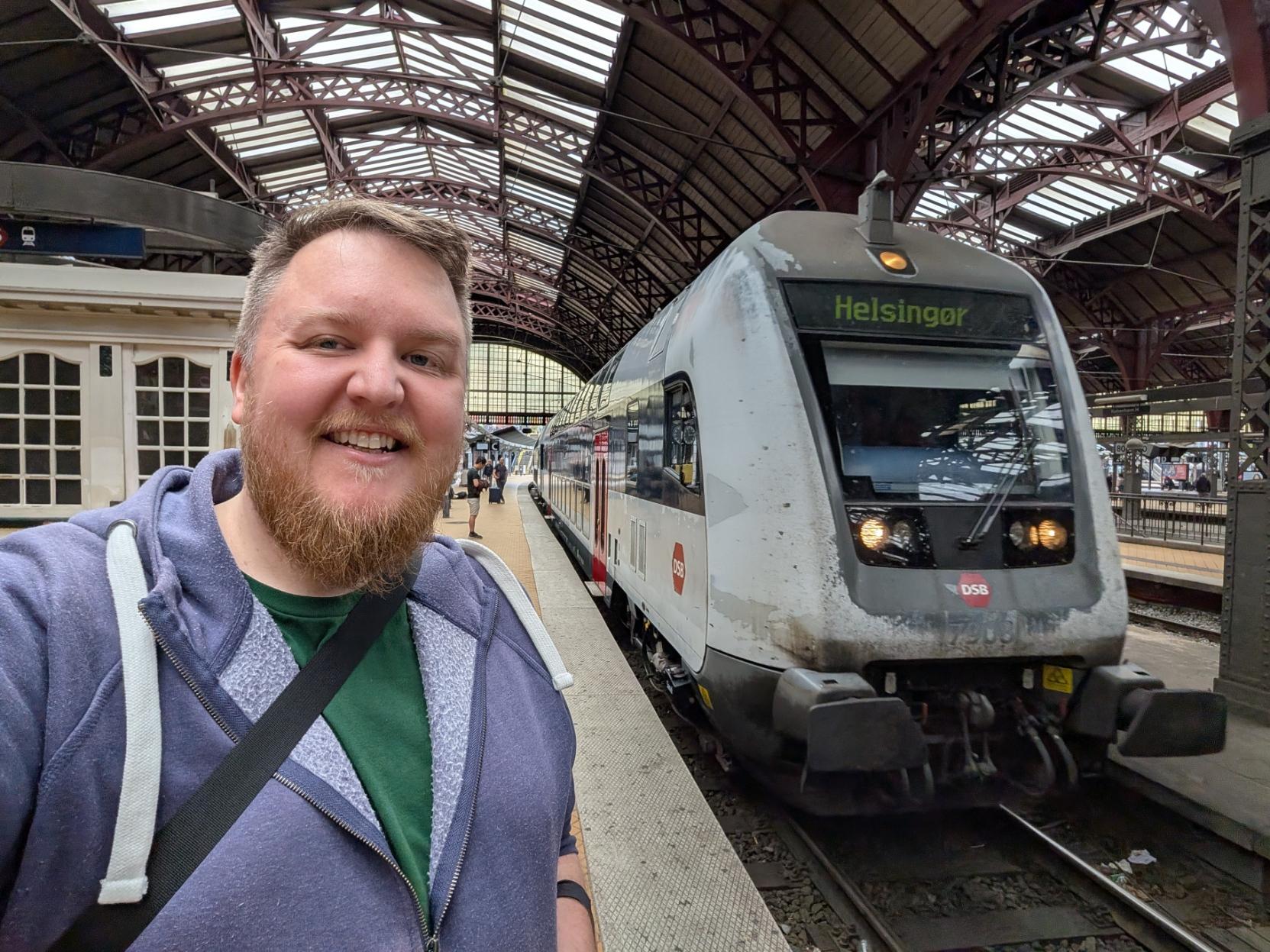 Ein selfie av ein smilande mann med skjegg og lilla hettegenser på ein perrong under den store bogehallen på Københavns Hovedbanegård. Til høgre for han står fronten på ei kvit og grå Bombardier dobbeltdrekar-styrevogn (DSB ABs). Toget har det raude DSB-logoen på snuten, og det digitale destinasjonsskiltet over frontruta lyser grønt med teksten «Helsingør». I bakgrunnen ser ein den opne og historiske stasjonsarkitekturen i tre og stål med bogetak, og nokre få andre reisande på perrongen.