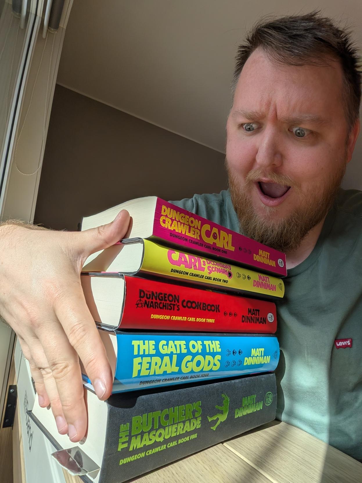 Ein selfie av ein mann med skjegg og salviegrøn t-skjorte som ser på ein bunke med bøker med eit overgira, sjokkert og storøygd uttrykk. Han held armen rundt ein stabel med fem tjukke fantasybøker i LitRPG-serien Dungeon Crawler Carl av Matt Dinniman, som ligg oppå eit lyst trebord. Bokryggene vender mot kameraet og viser titlane i sterke fargar nedover: den rosa «Dungeon Crawler Carl» (bok 1), den gule «Carl's Doomsday Scenario» (bok 2), den raude «The Dungeon Anarchist's Cookbook» (bok 3), den lyseblå «The Gate of the Feral Gods» (bok 4) og den svarte «The Butcher's Masquerade» (bok 5) med grøn skrift. I bakgrunnen strøymer det ljos inn frå eit vindauge til venstre.