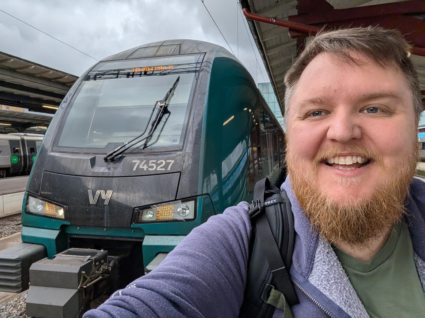 Ein selfie av ein breitt smilande mann med skjegg, grøn t-skjorte og lilla hettegenser på ein togperrong i dagslys. Til venstre står fronten på det same elektriske Vy-toget av typen Stadler FLIRT (Vy litra BM74) med settnummer «74527». Både det venstre og høgre markeringslyset nede lyser no i eit klart gult og kvitt skinn, og på den digitale destinasjonsskjermen øvst i frontruta kan ein skimte teksten «RE20 Oslo S». I bakgrunnen ser ein perrongtaket over spora under ein overskya, grå morgonhimmel, og delar av eit anna tog er synleg til venstre.