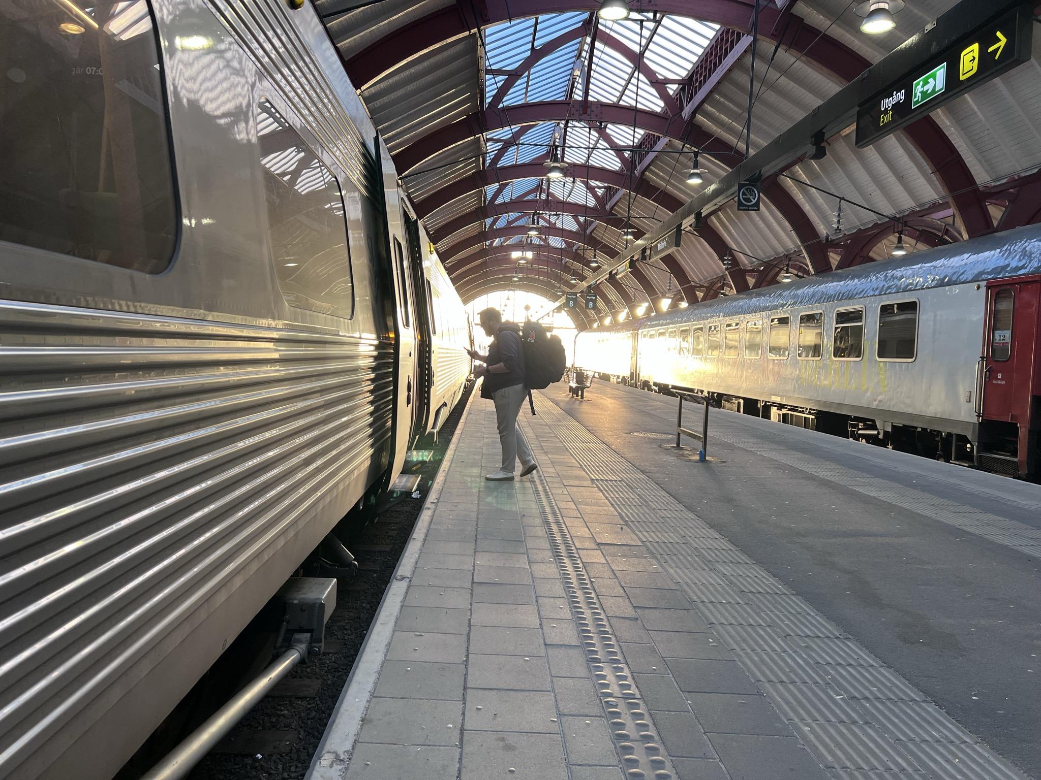 Bilete frå perrong på togstasjon. Til venstre i biletet er eit grått tog med dørane opne. Til høgre er eit grått tog med raude dørar. Taket er krummet med raude stålbjelkar og kvitt tak. Heilt i enden er sola så sterk at toga ikkje kan bli sett grunna det blendande ljoset.

Framføre sola er ein mann med stor ryggsekk på veg ombord i toget til venstre.