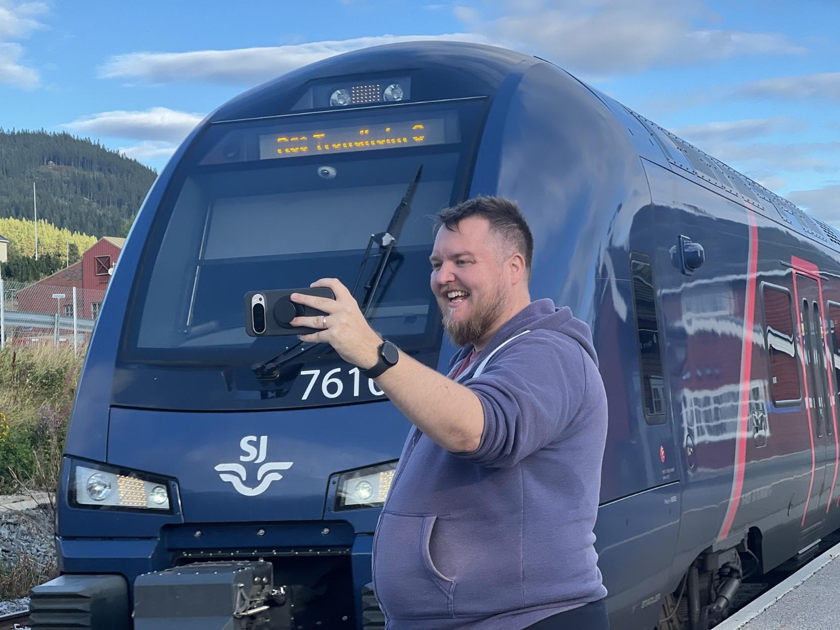 Ein mann med skjegg og lilla hettejakke står på perrongen på Alvdal stasjon og tek togselfie med mobilen i den løfta handa, breitt smilande med blikket på skjermen. Bak han fyller sida og fronten på eit mørkeblått bimodalt motorvognsett av typen Stadler FLIRT (SJ Nord litra BM76) heile biletet. Settnummeret «7610» og litt til står i kvit skrift under frontruta, delvis skjult av armen hans, og SJ-logoen med det vinga symbolet sit under. Destinasjonsskjermen over ruta lyser oransje, men er ikkje lesbar frå denne vinkelen. Ei raud stripe går langs vognsida, der den raude stasjonsbygningen speglar seg. I bakgrunnen til venstre ligg raudmåla hus og ein skogkledd ås under ein blå himmel med kvite skyer.

🚆 Tog: Type 76 · Litra: BM76 · Nr: 76101 · Stasjon: Alvdal · Bane: Rørosbanen · Selskap: SJ Nord