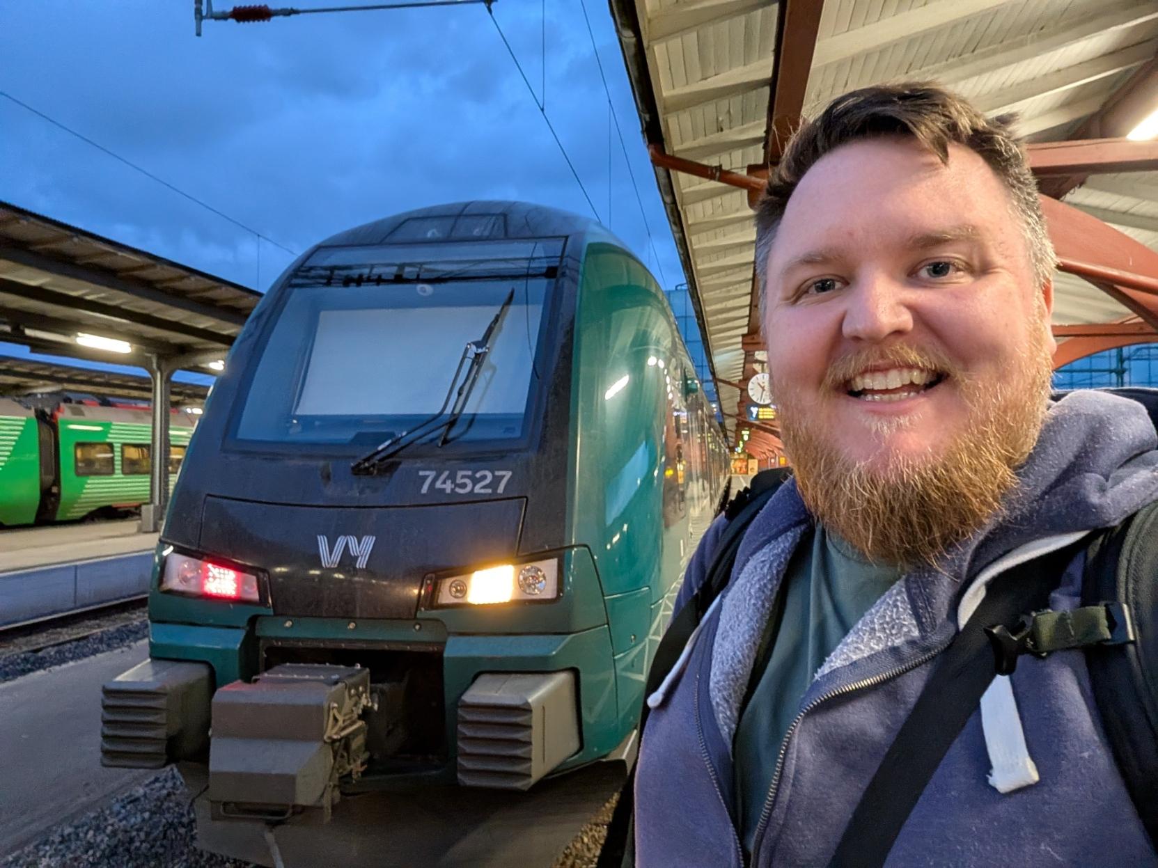 Ein selfie av ein breitt smilande mann med skjegg og lilla hettegenser på ein utandørs togperrong om kvelden (Göteborg Centralstation). Til venstre står fronten på eit grønt og mørkegrått elektrisk tog av typen Stadler FLIRT (Vy litra BM74) parkert for natta. Togfronten er merka med Vy sin kvite logo og har settnummeret «74527» under frontruta. Det venstre markeringslyset lyser raudt, medan det høgre lyser gult. I bakgrunnen til venstre kan ein sjå eit anna grønt tog parkert ved naboperrongen, og over perrongtaka breier ein mørkeblå kveldshimmel seg ut.