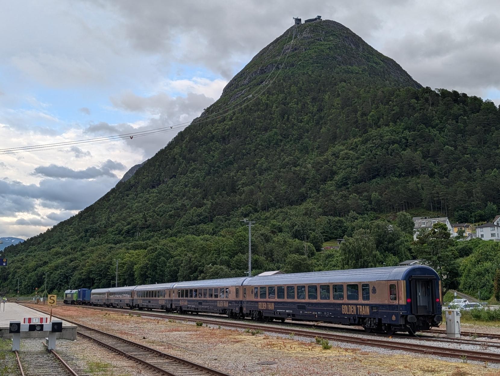 Eit oversiktsbilete frå stasjonsområdet på Åndalsnes stasjon, der turisttoget The Golden Train står parkert på eit sidespor. Togsettet strekkjer seg bortover skjenegangen med sine særprega mørkeblå passasjervogner som har eit breitt, gyllent gullfelt langs vindaugsrekkja og teksten «THE GOLDEN TRAIN» på vognsidene. I fronten av togstamma kan ein skimte eit grønt og kvitt diesellokomotiv. Rett bak toget reiser det mektige og skogkledde fjellet Nesaksla seg steilt opp mot ein skyete himmel, og på toppen kan ein sjå fjellstova Eggen. Strekkjer ein auga langs kabelen frå stasjonen og oppover fjellsida, kan ein sjå Romsdalsgondolen på veg opp mot toppen.